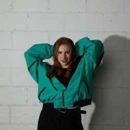 Kub1k - Portret uśmiechniętej kobiety z rudymi włosami, w turkusowej kurtce retro, pozującej na tle ściany z białych cegieł.