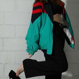 Kub1k - Rudowłosa modelka w czarnych spodniach i sportowej kurtce, trzymająca czarny lakierowany szpilkę przy stopie na tle białej ceglanej ściany.