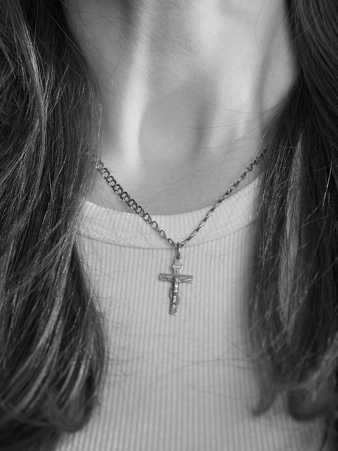 Czarno-białe zbliżenie na szyję kobiety z długimi włosami i srebrnym łańcuszkiem z krzyżykiem na białej bluzce z prążkami.