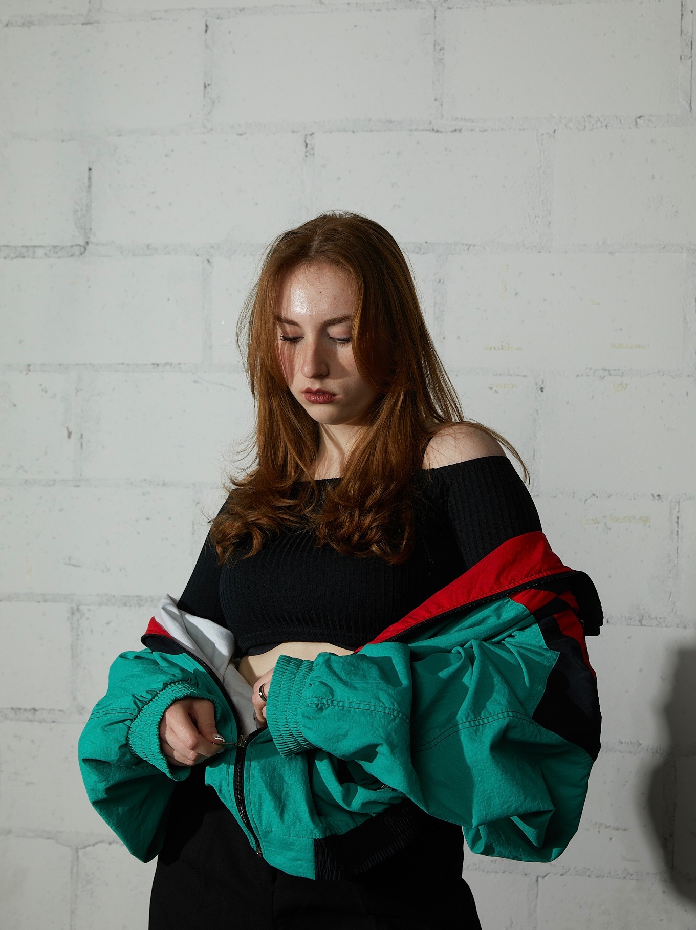 Młoda kobieta z rudymi włosami, ubrana w czarny top z odkrytymi ramionami, zapinająca zielono-czerwono-białą kurtkę na tle białej, ceglanej ściany.