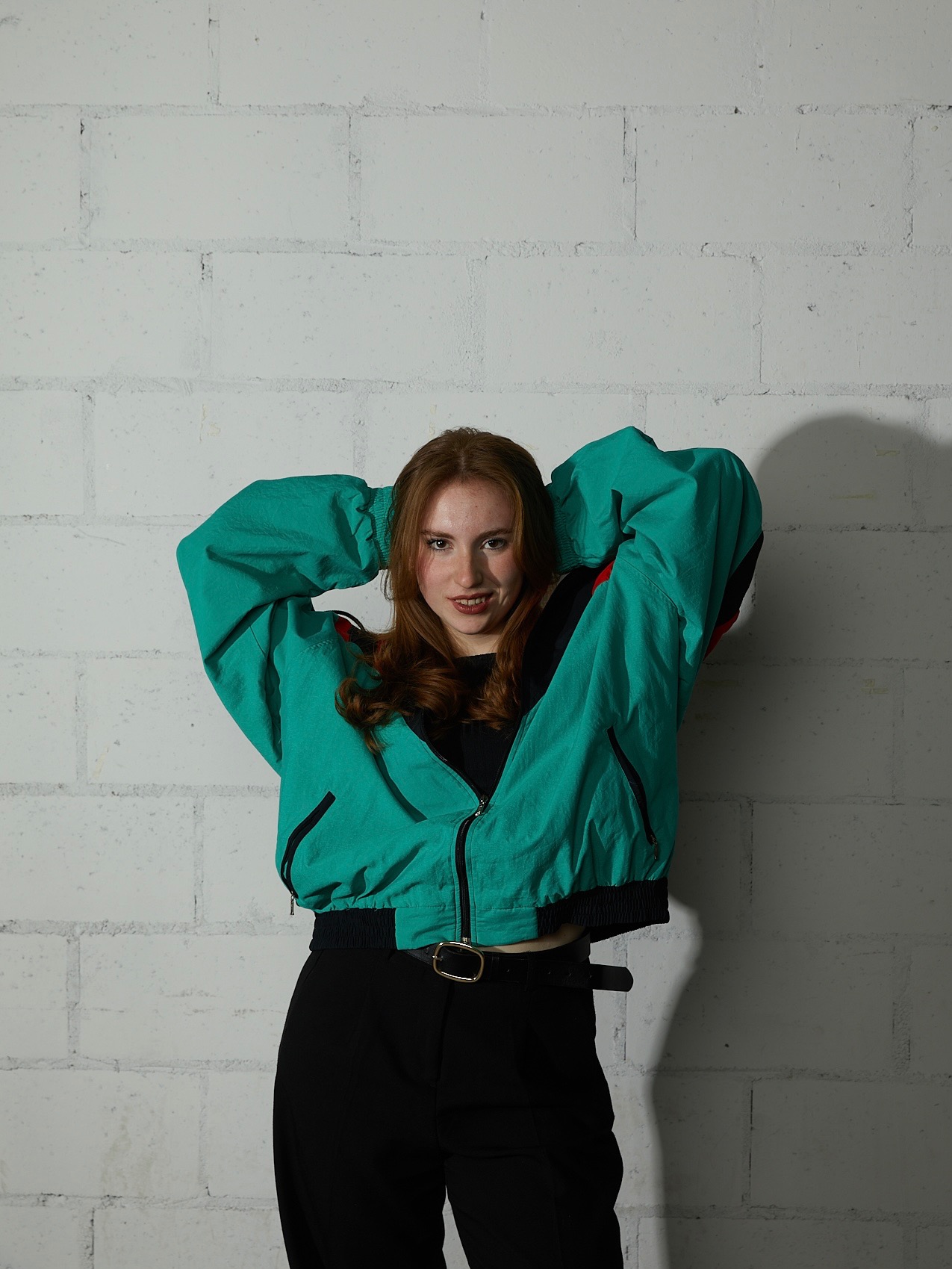 Portret uśmiechniętej kobiety z rudymi włosami, w turkusowej kurtce retro, pozującej na tle ściany z białych cegieł.