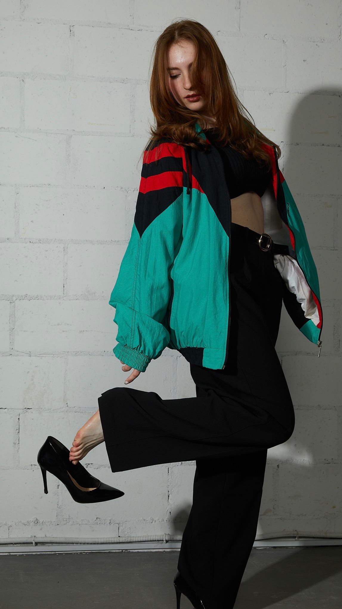 Rudowłosa modelka w czarnych spodniach i sportowej kurtce, trzymająca czarny lakierowany szpilkę przy stopie na tle białej ceglanej ściany.