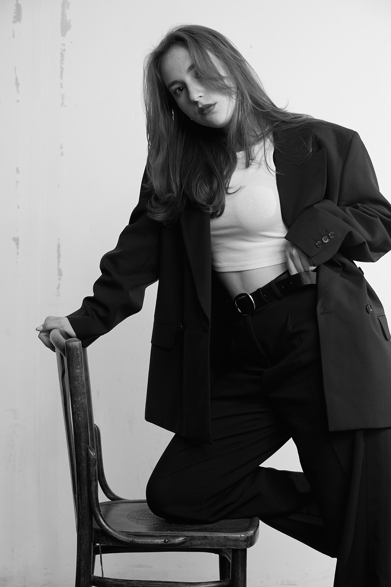 Czarno-białe zdjęcie kobiety w oversize garniturze, opierającej się o stare krzesło, z ręką w kieszeni spodni i zamyślonym wyrazem twarzy.