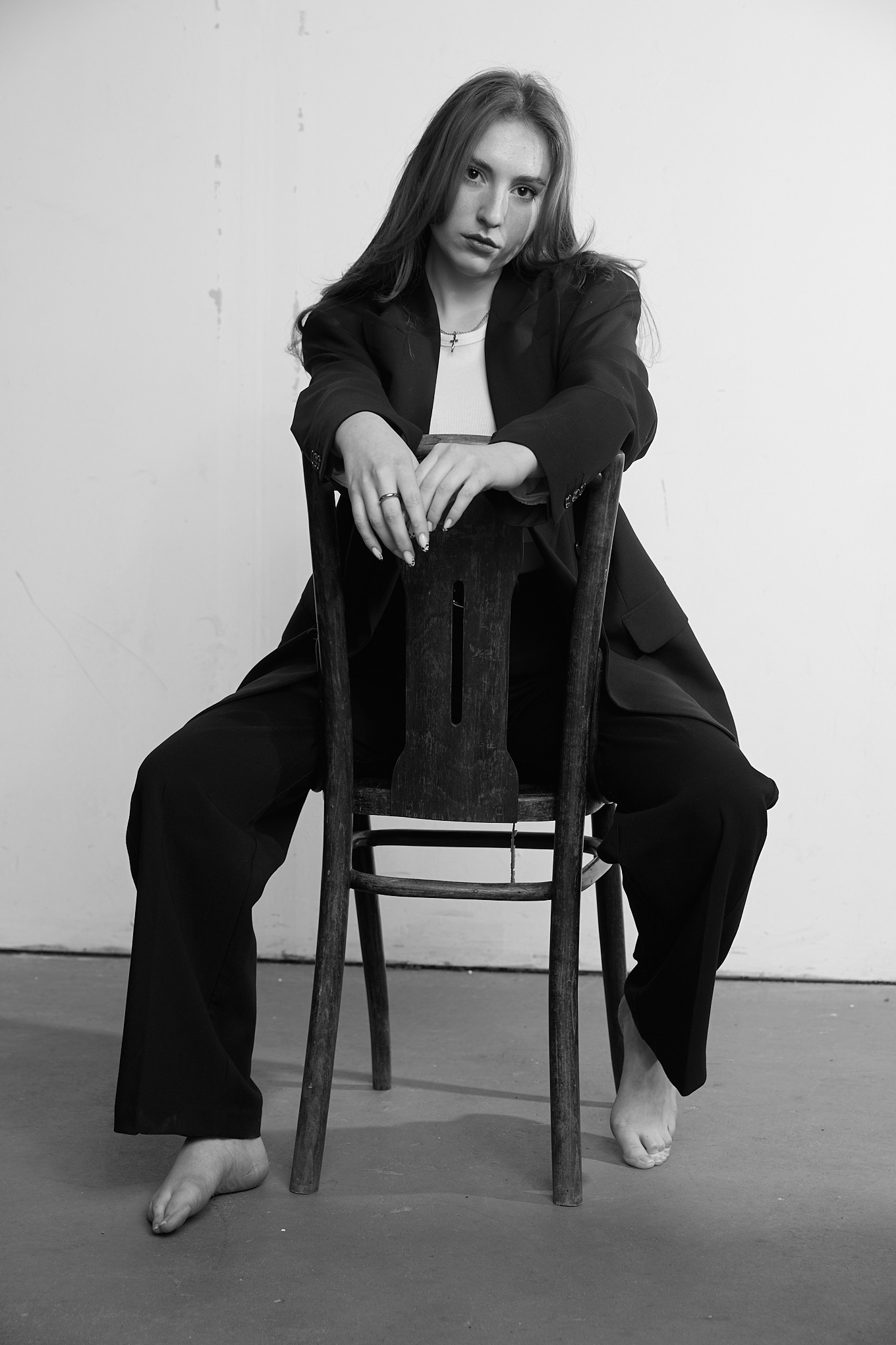 Czarno-białe zdjęcie kobiety siedzącej na krześle w oversize'owym garniturze, boso, z rękami opartymi na oparciu krzesła, patrzącej w obiektyw z melancholijnym wyrazem twarzy.