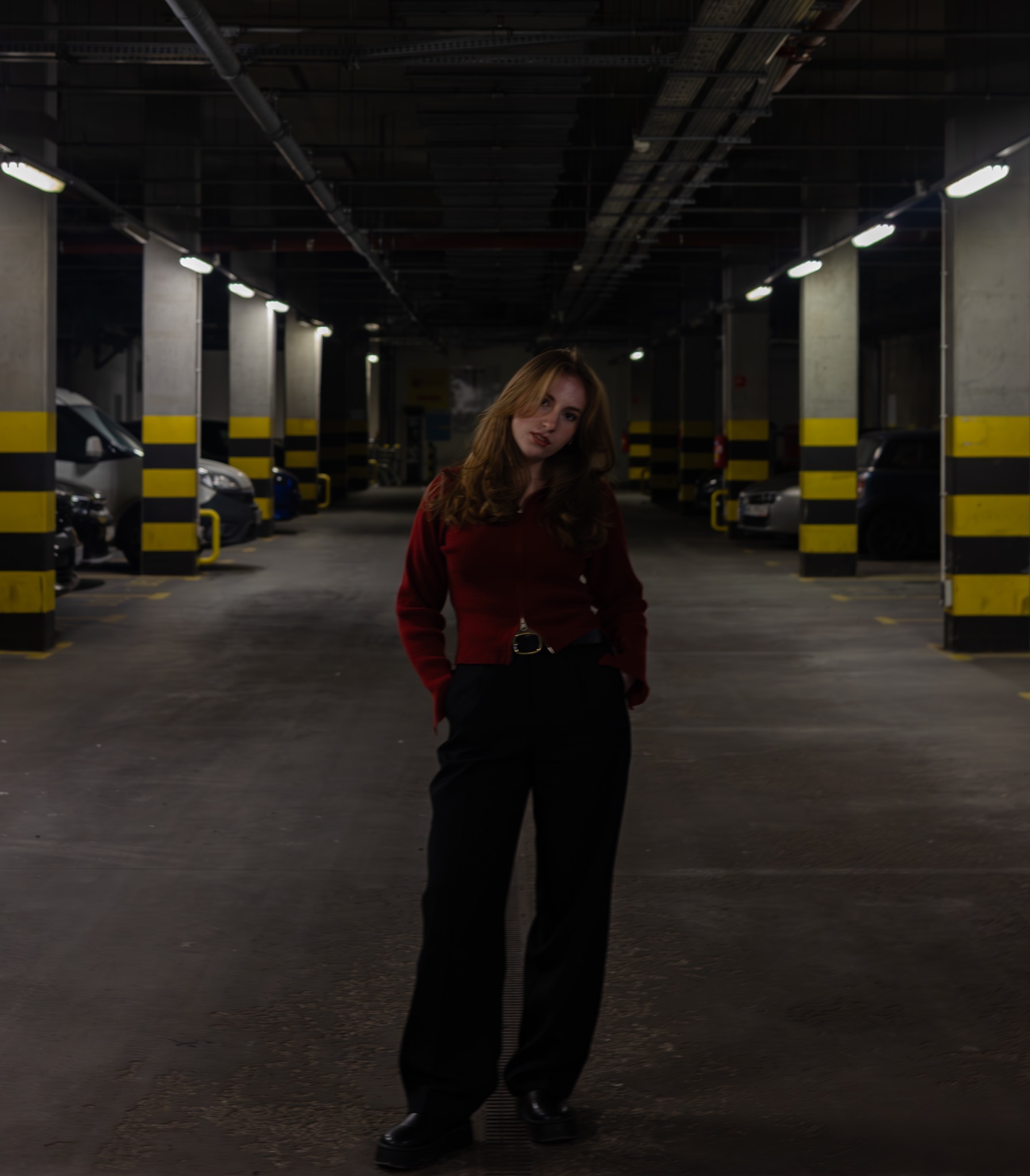 Portret kobiety w czerwonym swetrze i czarnych spodniach na tle parkingu podziemnego z rzędami kolumn w żółto-czarne pasy i zaparkowanymi samochodami.