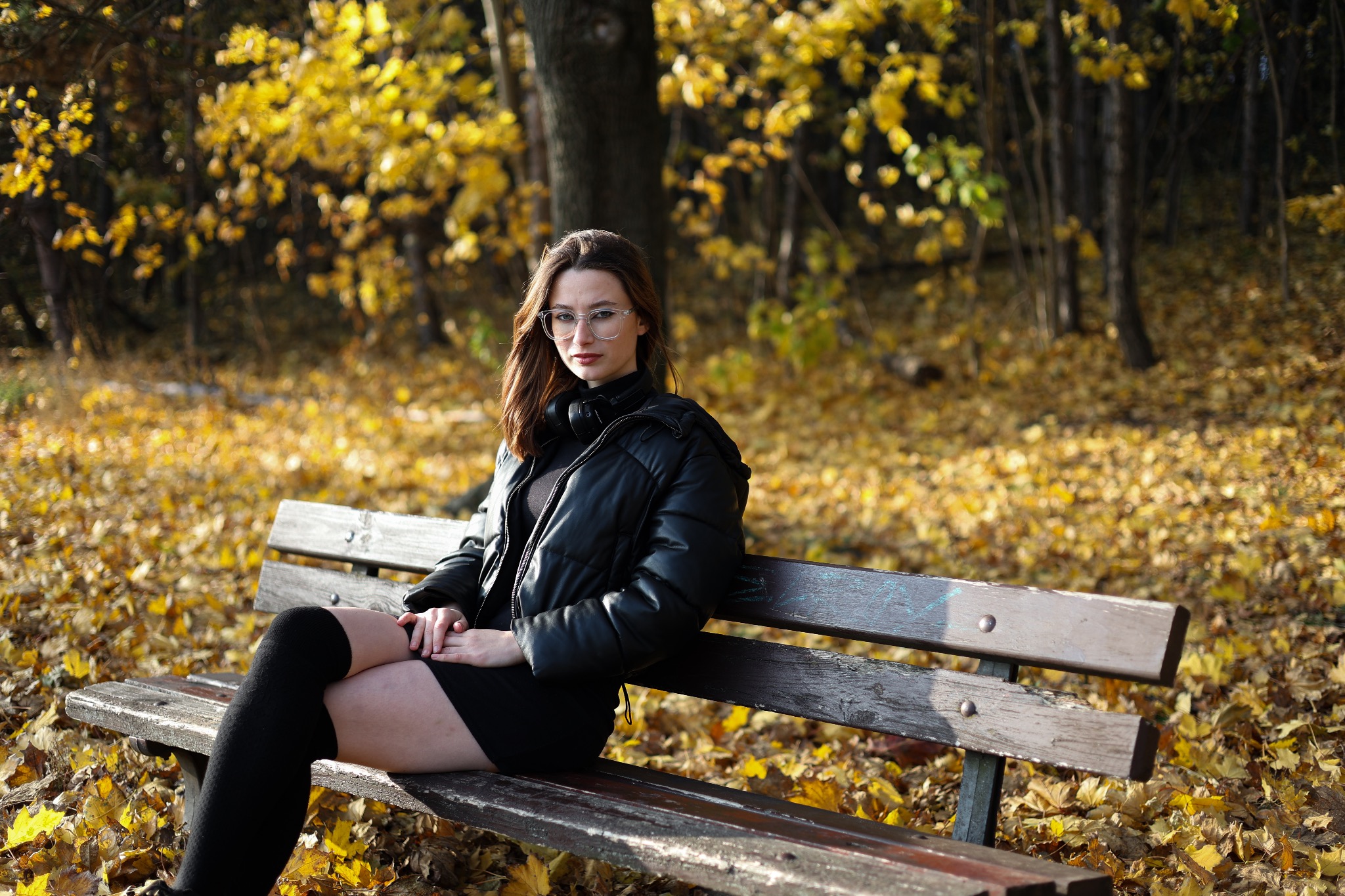 Kobieta w okularach siedzi na ławce w parku, ubrana w czarną kurtkę i podkolanówki, jesienne liście w tle.
