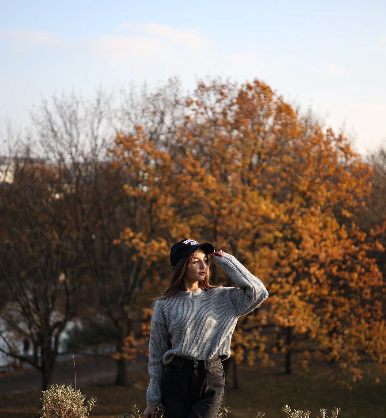 Młoda kobieta w szarym swetrze i czarnej czapce z daszkiem pozuje na tle jesiennych drzew z pomarańczowymi liśćmi, trzymając ręką daszek czapki i patrząc w bok.