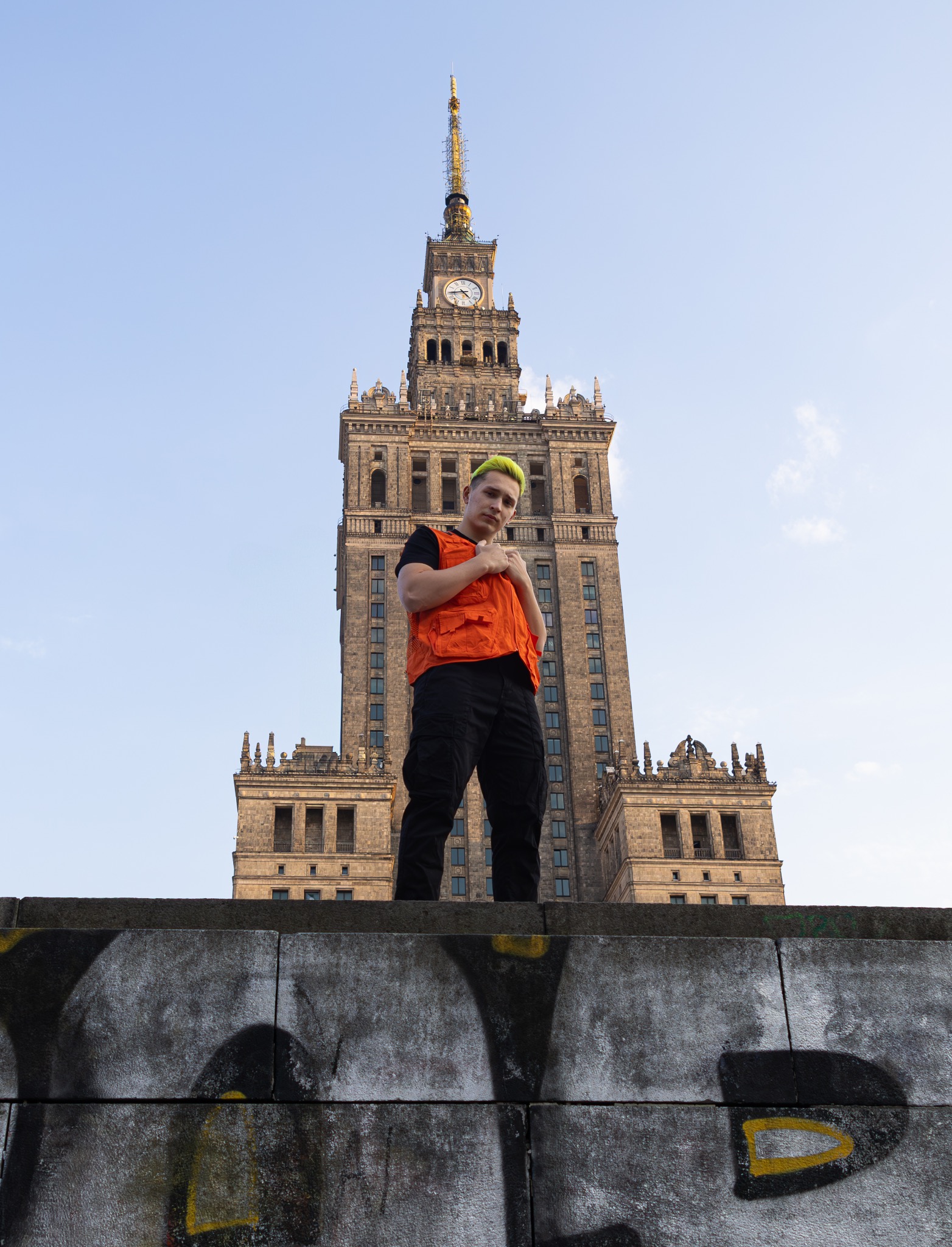 Młody mężczyzna z neonowo-zielonymi włosami w pomarańczowej kamizelce pozuje na tle Pałacu Kultury i Nauki w Warszawie, stojąc na betonowym murku z graffiti.