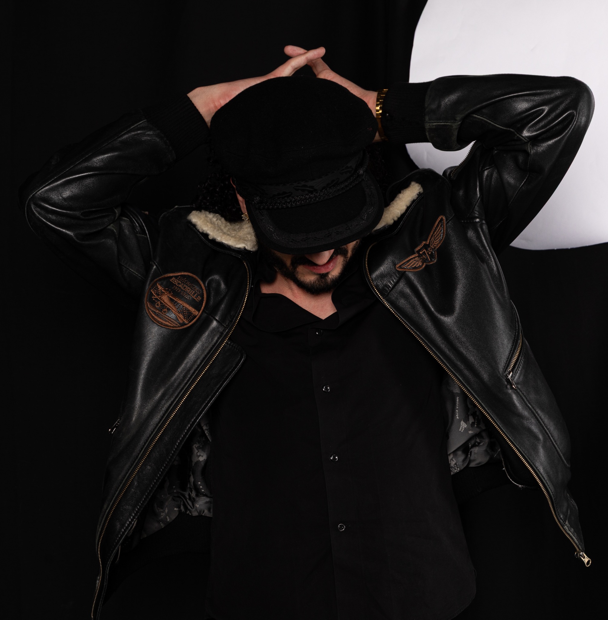 Mężczyzna w czarnej skórzanej kurtce z naszywkami i czarnym kapeluszu, ręce uniesione nad głową, na czarnym tle.