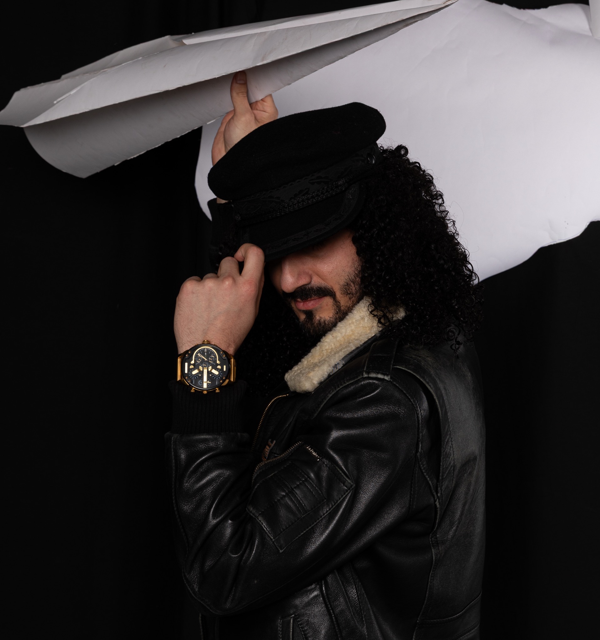 Mężczyzna w czarnej skórzanej kurtce i czapce kapitańskiej pozuje na czarnym tle, z dłonią na czapce i dużym złotym zegarkiem na nadgarstku, oświetlony od góry białym reflektorem.
