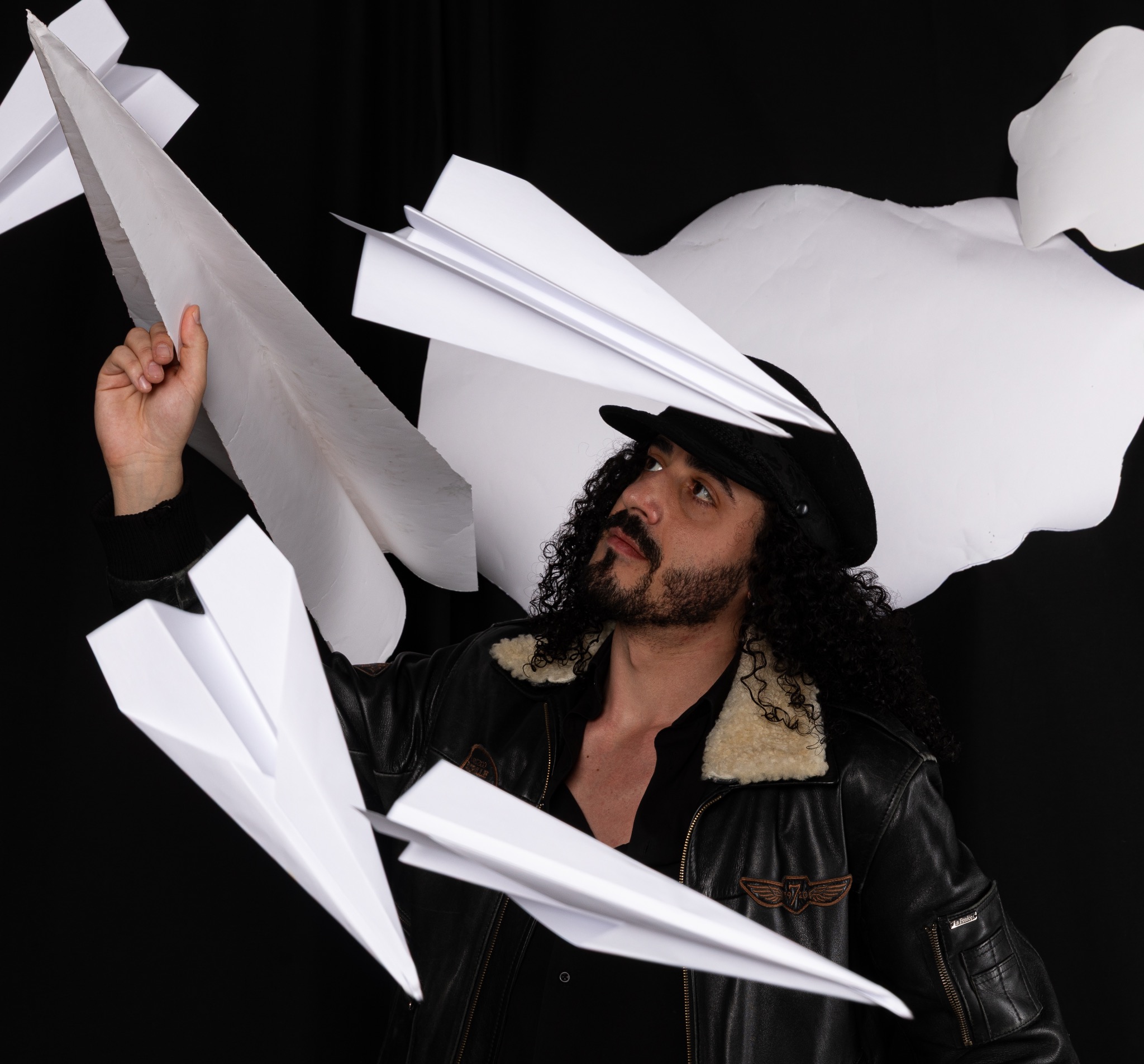 Mężczyzna w skórzanej kurtce i kapeluszu otoczony papierowymi samolotami na czarnym tle, spoglądający w górę.