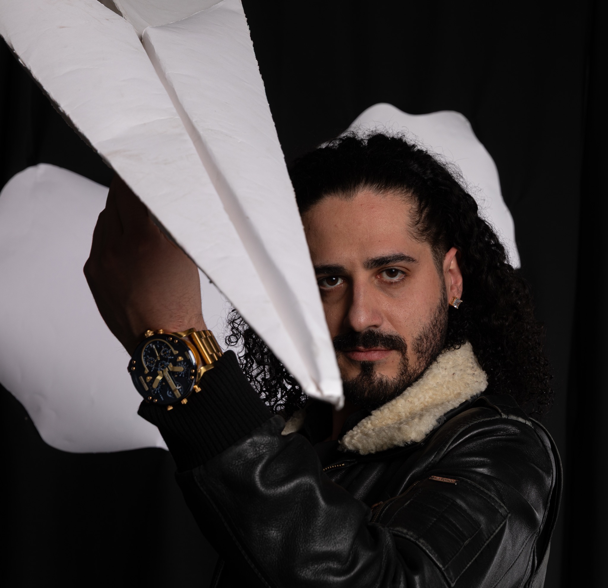 Portret mężczyzny z kręconymi włosami i brodą, w skórzanej kurtce z kożuszkiem, trzymającego papierowy samolot, na czarnym tle.