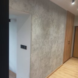 PROF-REMONT-EXPRES BOHDAN KOMARYNSKYI - Ściana z dekoracyjnym tynkiem w odcieniach szarości, sąsiadująca z drewnianą szafą w zabudowie i białym fragmentem ściany z czarnymi włącznikami światła, na podłodze panele imitujące drewno.