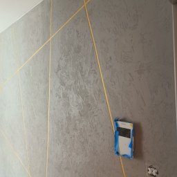 PROF-REMONT-EXPRES BOHDAN KOMARYNSKYI - Szara ściana z dekoracyjną fakturą i złotymi, ukośnymi liniami, zabezpieczone gniazdko elektryczne taśmą malarską.