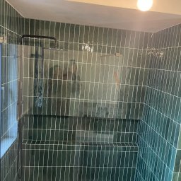 PROF-REMONT-EXPRES BOHDAN KOMARYNSKYI - Kabina prysznicowa wyłożona pionowymi, ciemnozielonymi płytkami z białymi fugami, widoczna szklana ściana i deszczownica, sufit z gołą żarówką.