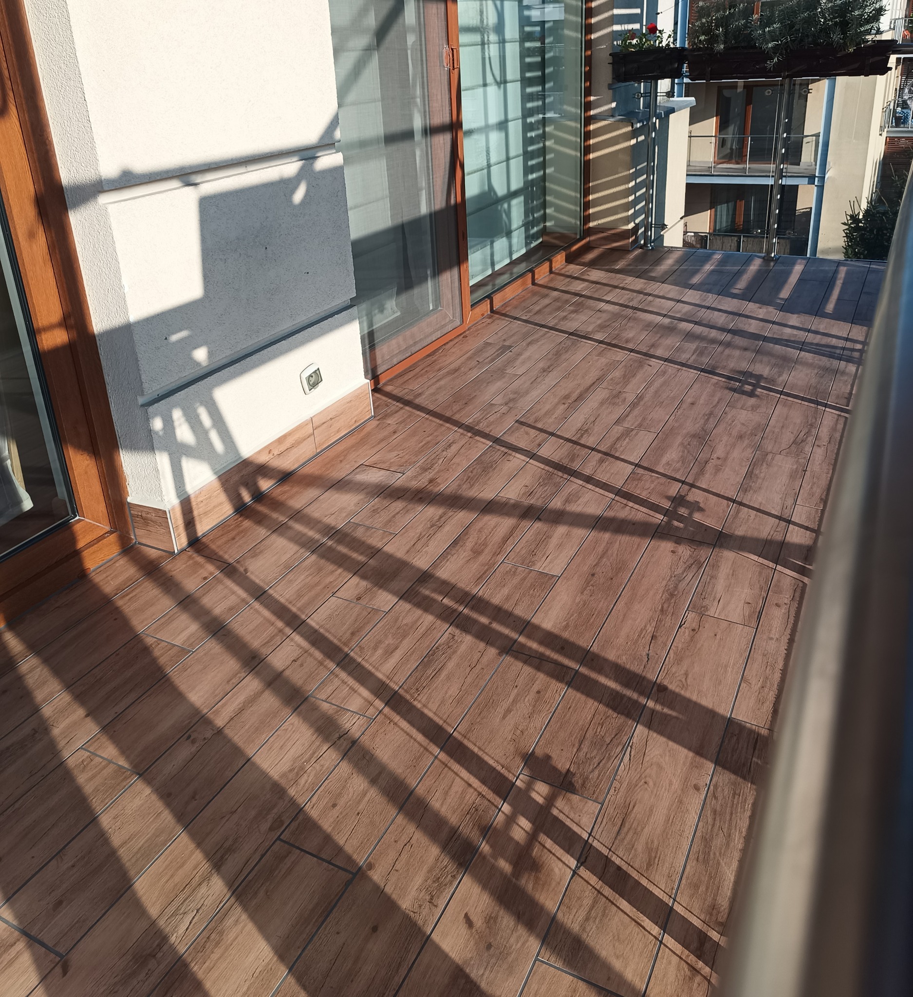 Balkon z podłogą wyłożoną płytkami imitującymi drewno, widoczne przesuwne drzwi balkonowe i cień rzucany przez balustradę.
