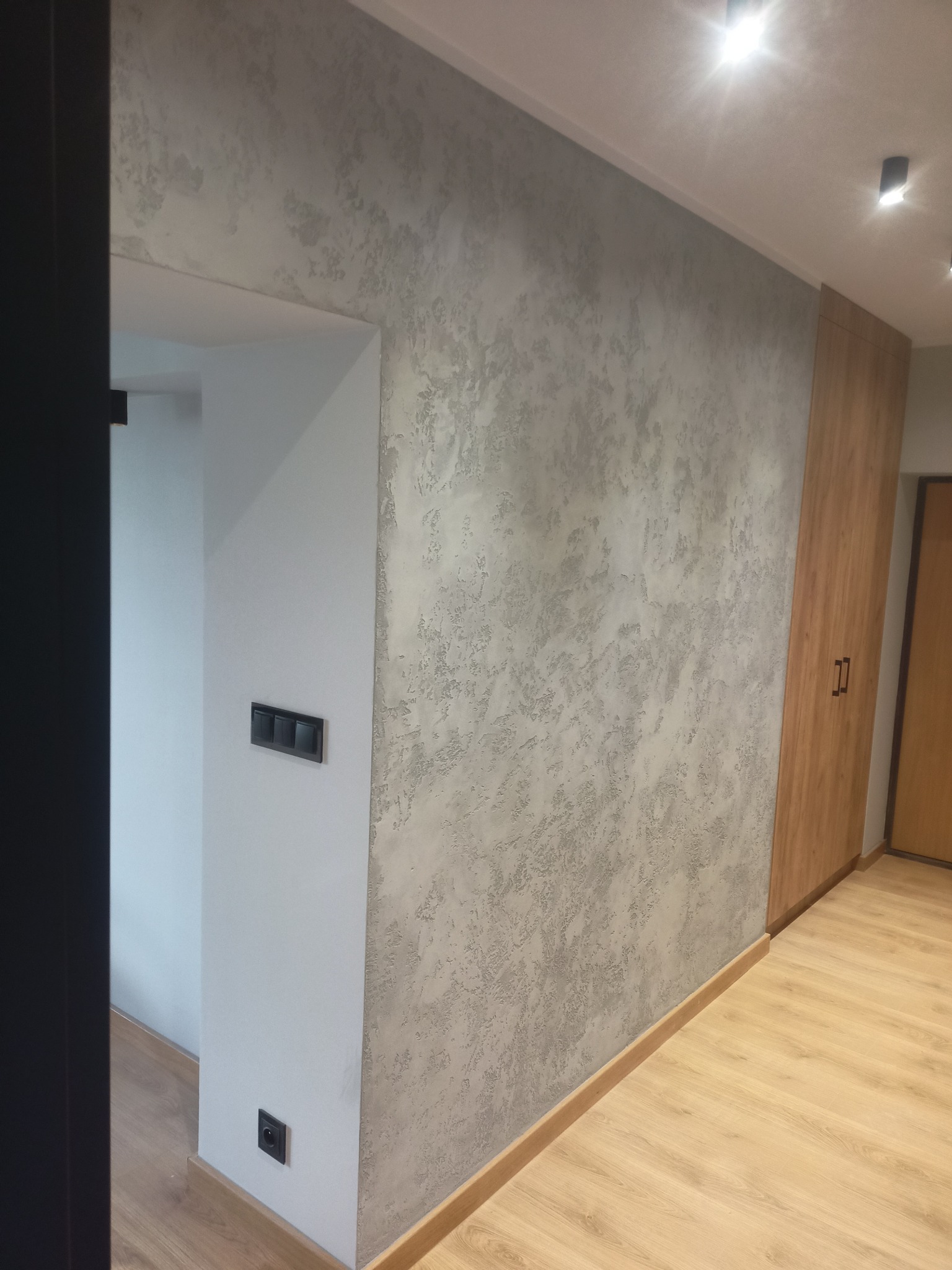 Ściana z dekoracyjnym tynkiem w odcieniach szarości, sąsiadująca z drewnianą szafą w zabudowie i białym fragmentem ściany z czarnymi włącznikami światła, na podłodze panele imitujące drewno.