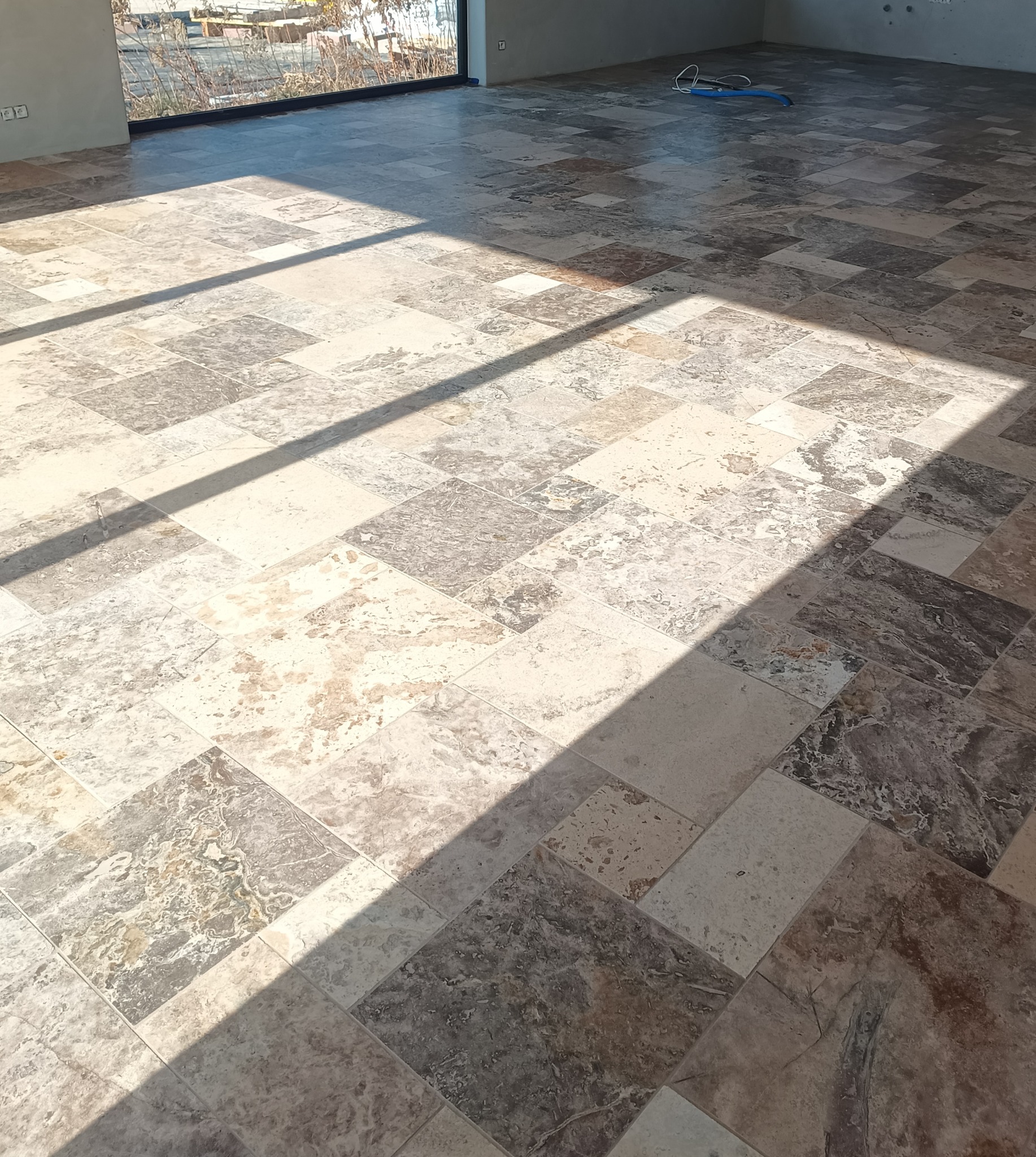 Ułożona podłoga z płytek imitujących kamień w odcieniach beżu i szarości, z widocznym fragmentem okna i wpadającym światłem słonecznym.