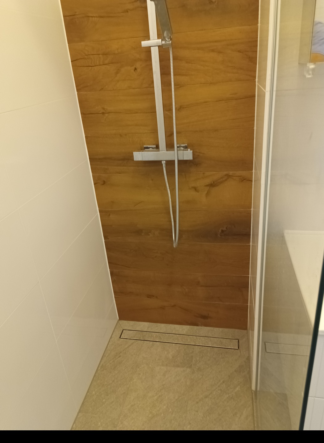 Kabina prysznicowa z drewnopodobnymi płytkami na ścianie, odpływ liniowy w podłodze i białe płytki na sąsiedniej ścianie.