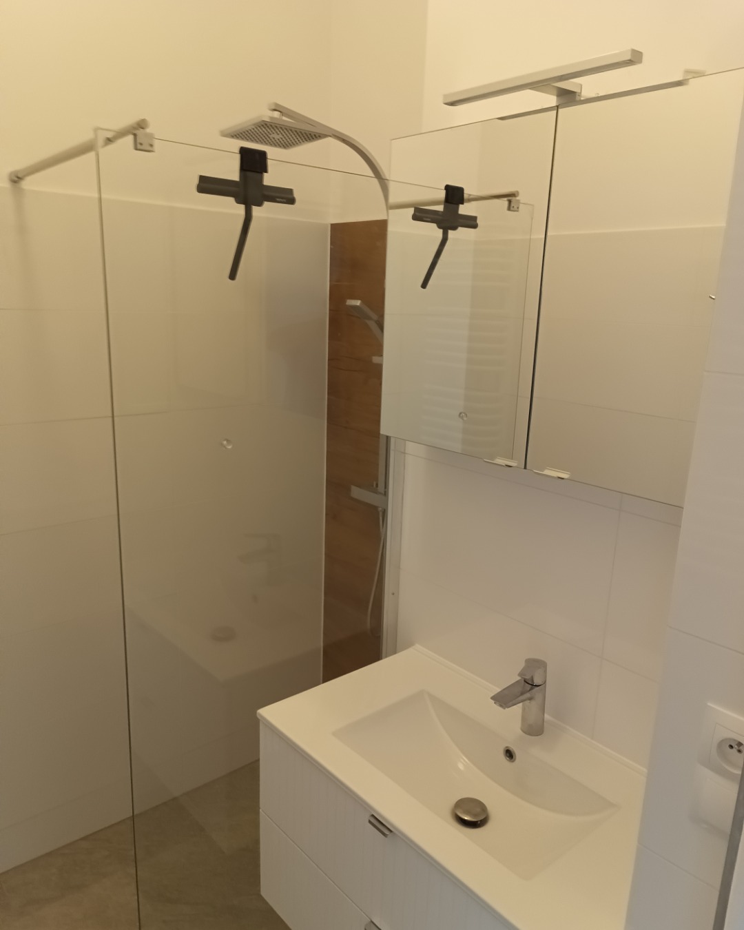 Nowoczesna kabina prysznicowa ze szklaną ścianką i deszczownicą w minimalistycznej łazience w Warszawie, widok na umywalkę z szafką i duże lustro.