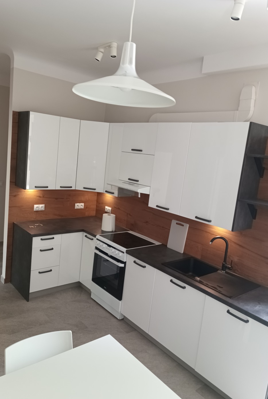 Nowoczesna kuchnia z białymi szafkami i ciemnym blatem, drewnopodobna ściana, oświetlenie punktowe i duża lampa wisząca nad stołem.