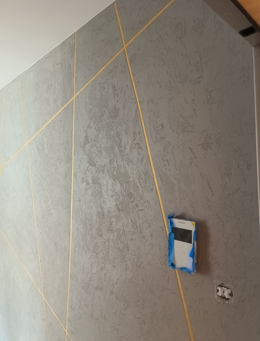 Szara ściana z dekoracyjną fakturą i złotymi, ukośnymi liniami, zabezpieczone gniazdko elektryczne taśmą malarską.