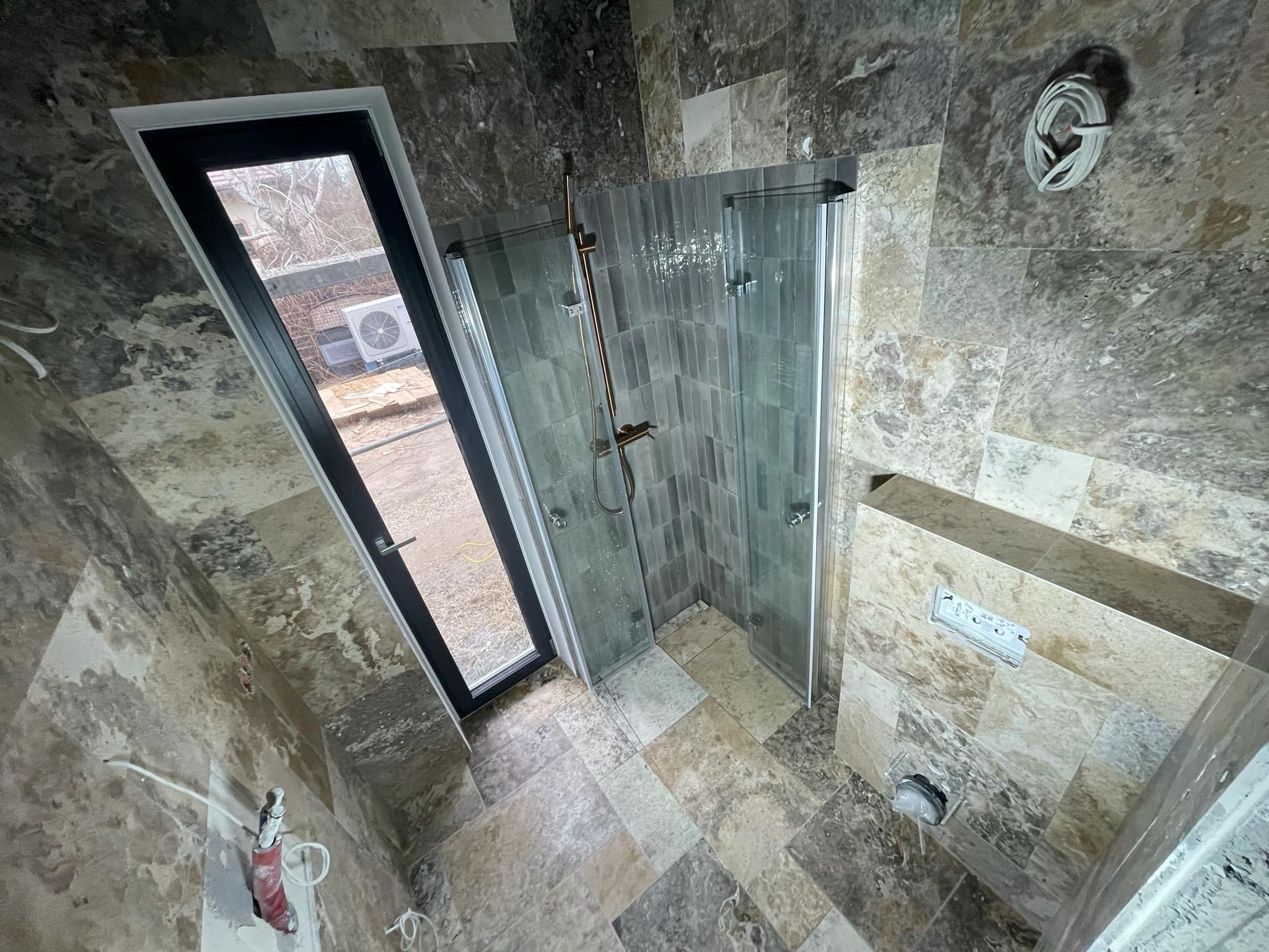 Wnętrze łazienki w trakcie prac wykończeniowych; widoczna kabina prysznicowa z elementami w kolorze złota, okno z czarną ramą, oraz ściany i podłoga wyłożone płytkami imitującymi kamień...