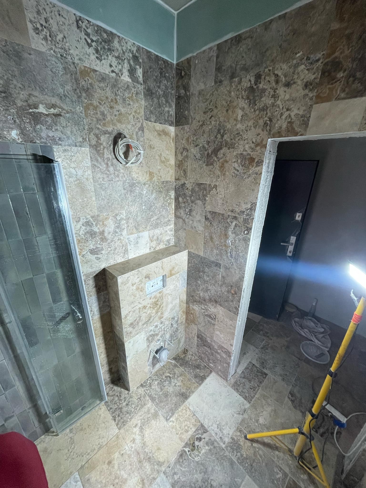 Wnętrze łazienki w trakcie remontu, widoczne płytki imitujące kamień na ścianach i podłodze, częściowo zamontowana kabina prysznicowa, stelaż podtynkowy WC oraz przewody elektryczne wystające ze...