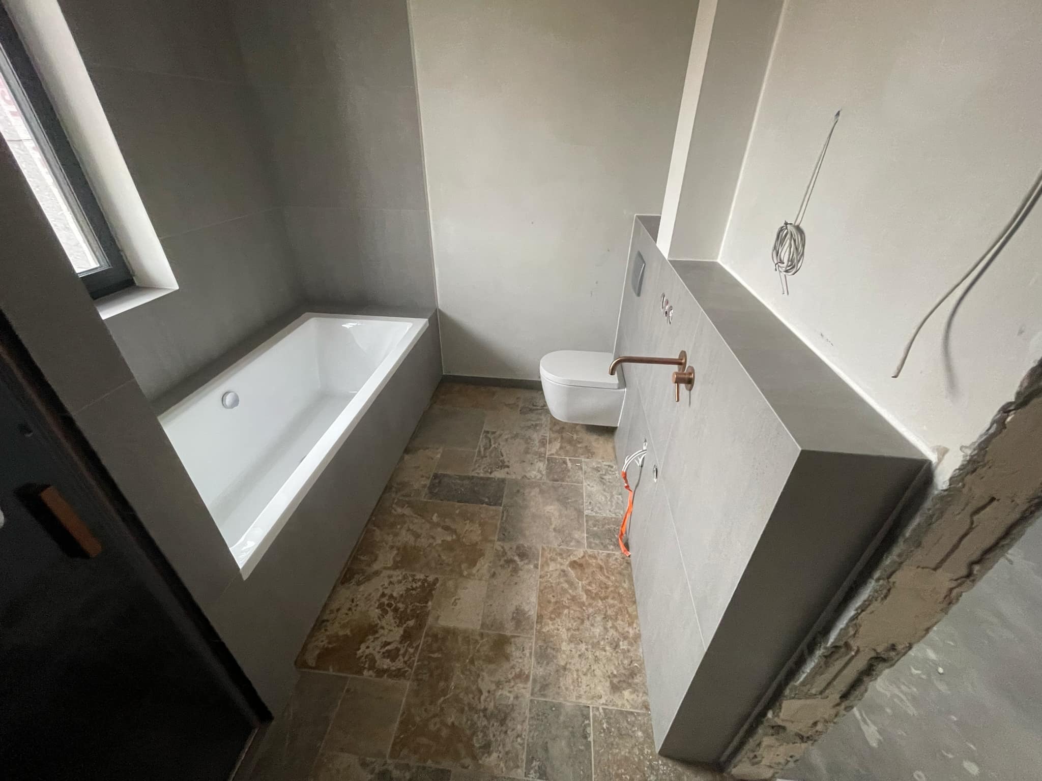 Wnętrze łazienki w trakcie wykańczania: biała wanna i toaleta, szare płytki na ścianach, podłoga z płytek imitujących kamień, wystające przewody elektryczne, widoczne ślady prac wykończeniowych.