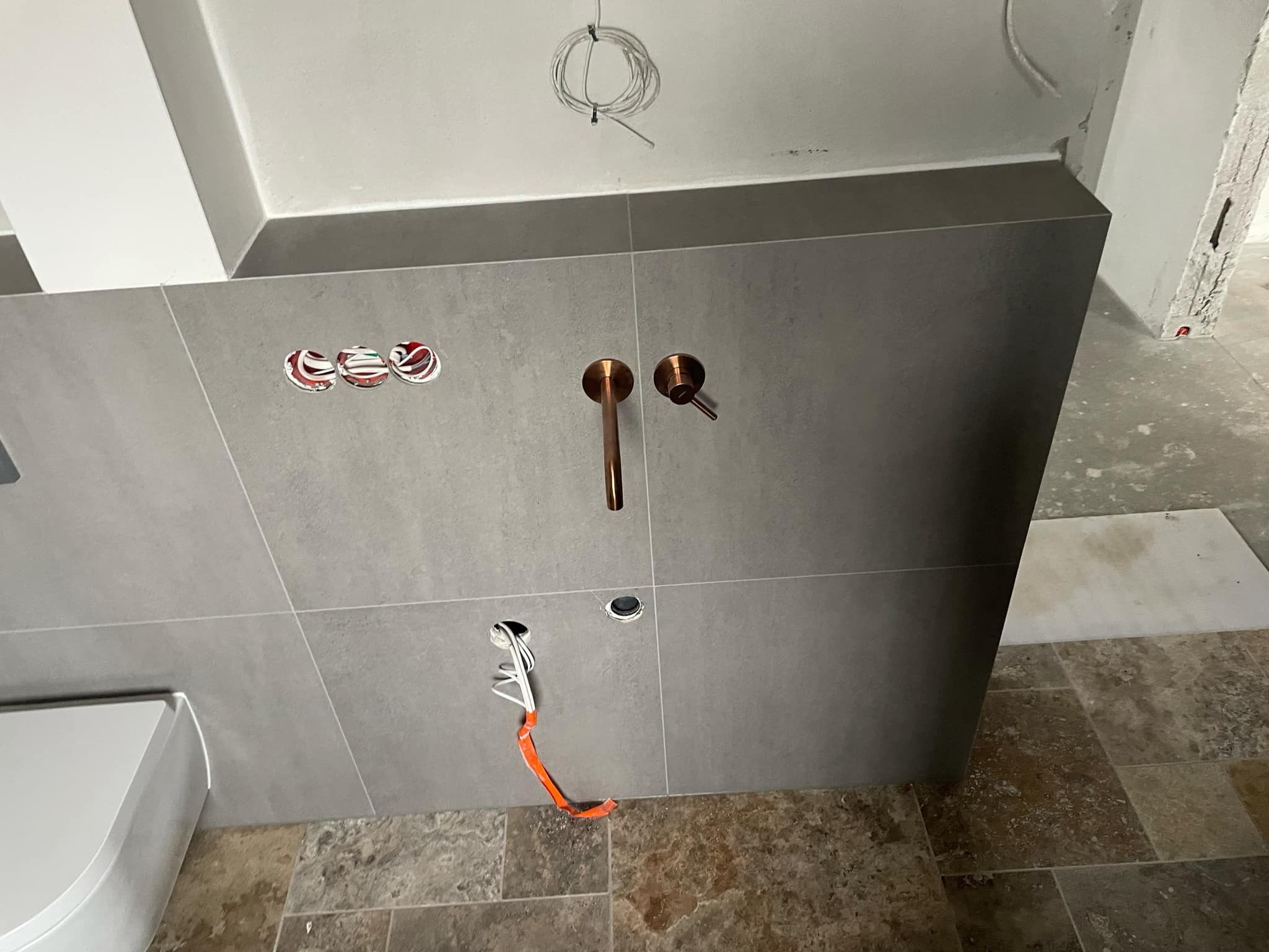 Szara ściana w trakcie montażu armatury łazienkowej z miedzianymi bateriami, widoczne wystające kable i otwory w płytkach, podłoga wyłożona płytkami imitującymi kamień.