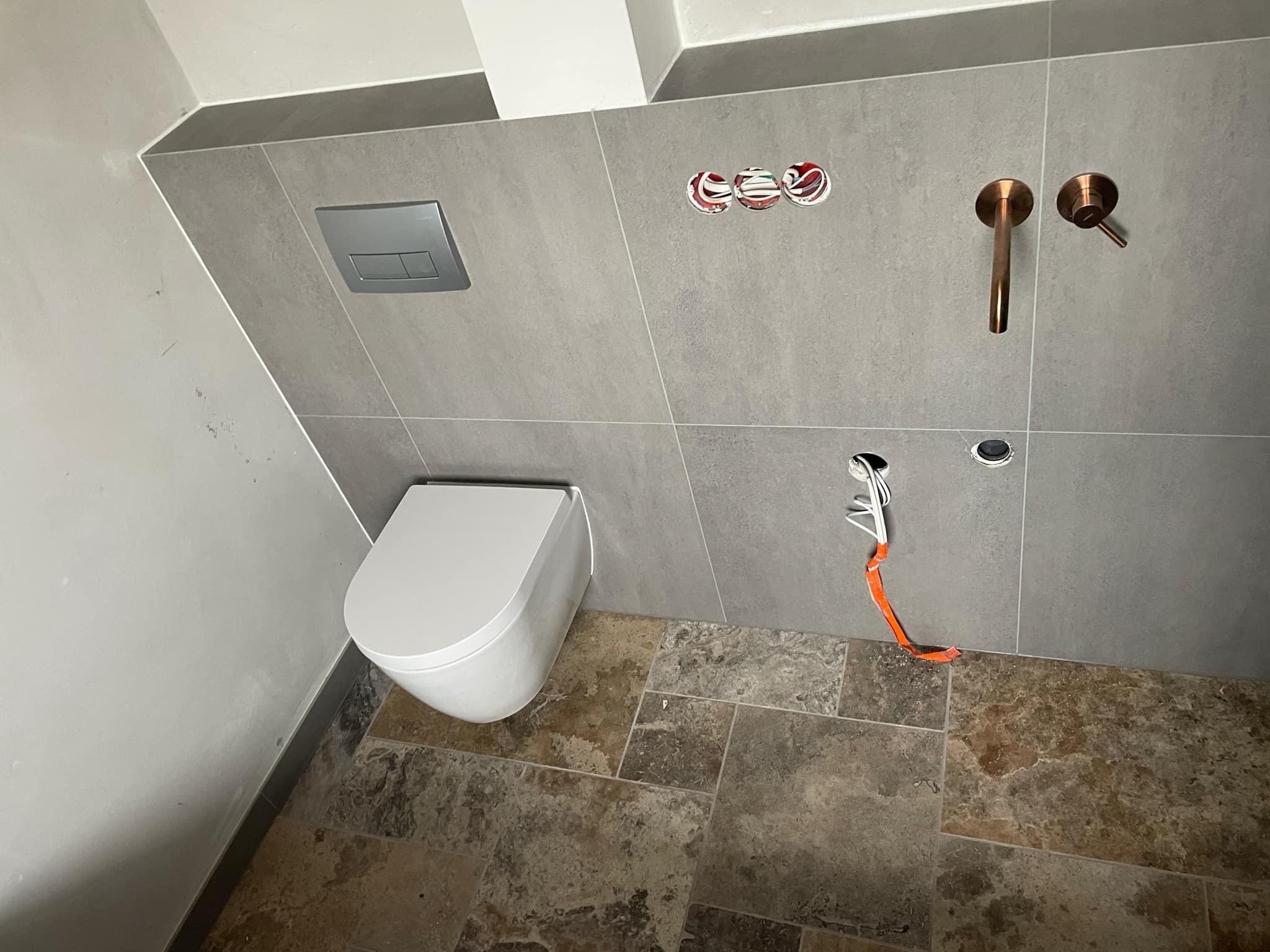 Wisząca toaleta z szarą płytką ścienną, miedziana armatura, instalacja elektryczna wystająca ze ściany i brązowa płytka podłogowa.