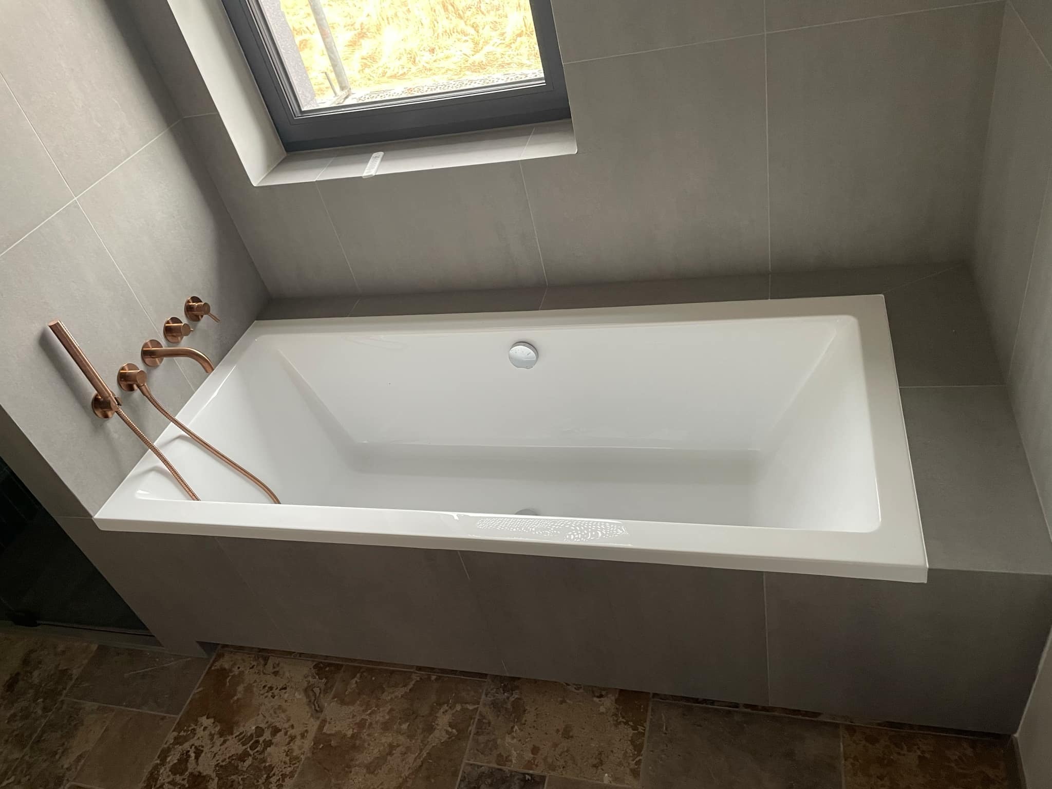 Nowoczesna, prostokątna wanna w obudowie z szarych płytek, z miedzianą armaturą i oknem dachowym.