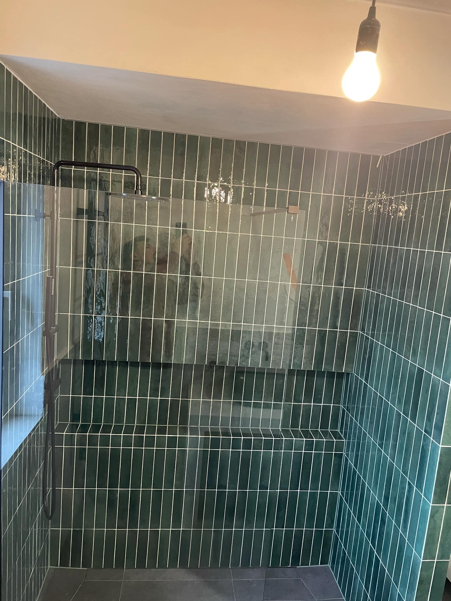 Kabina prysznicowa wyłożona pionowymi, ciemnozielonymi płytkami z białymi fugami, widoczna szklana ściana i deszczownica, sufit z gołą żarówką.
