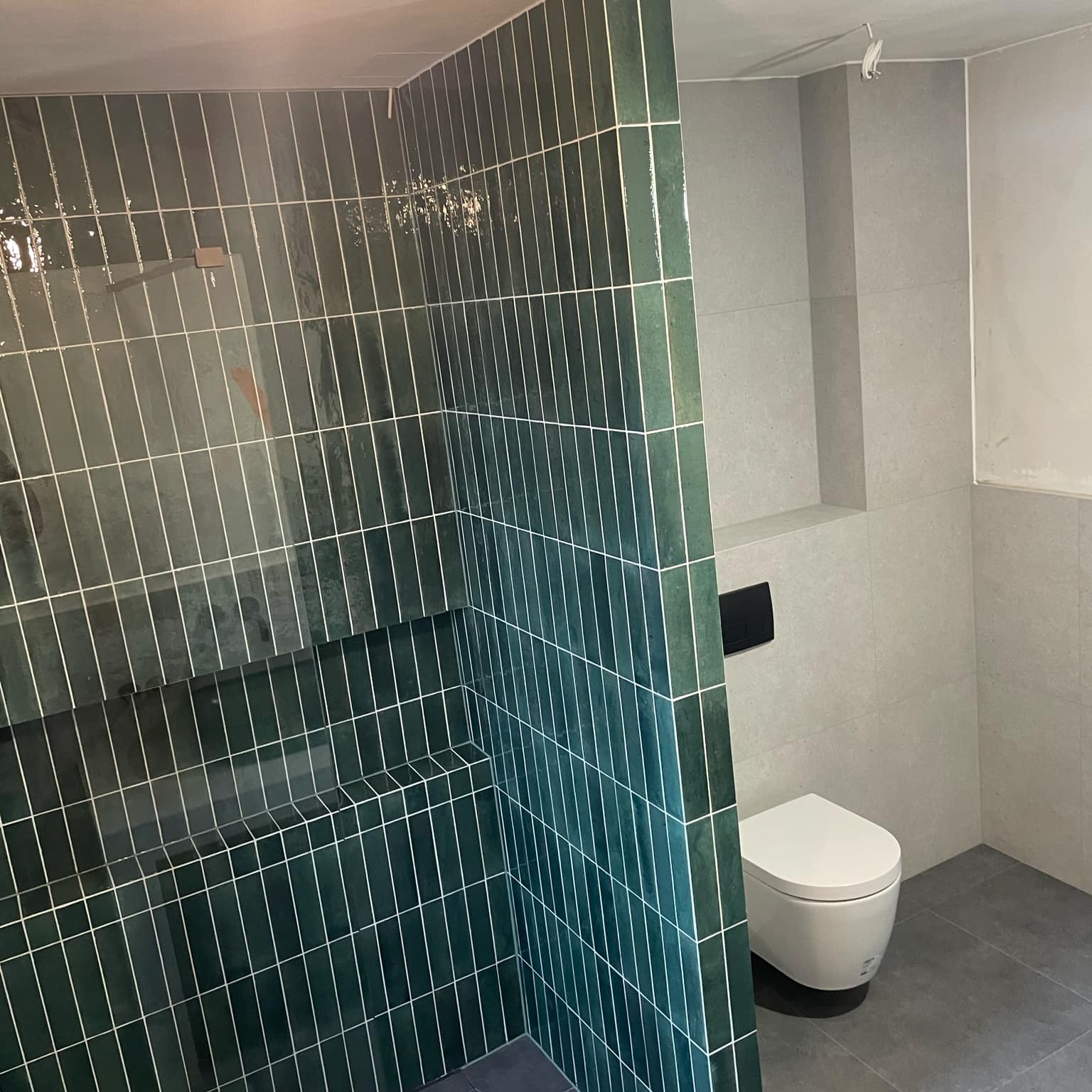 Nowoczesna łazienka z kabiną prysznicową wyłożoną pionowymi, zielonymi płytkami o błyszczącej powierzchni, kontrastującymi z szarymi płytkami na pozostałych ścianach i podłodze; widoczna...