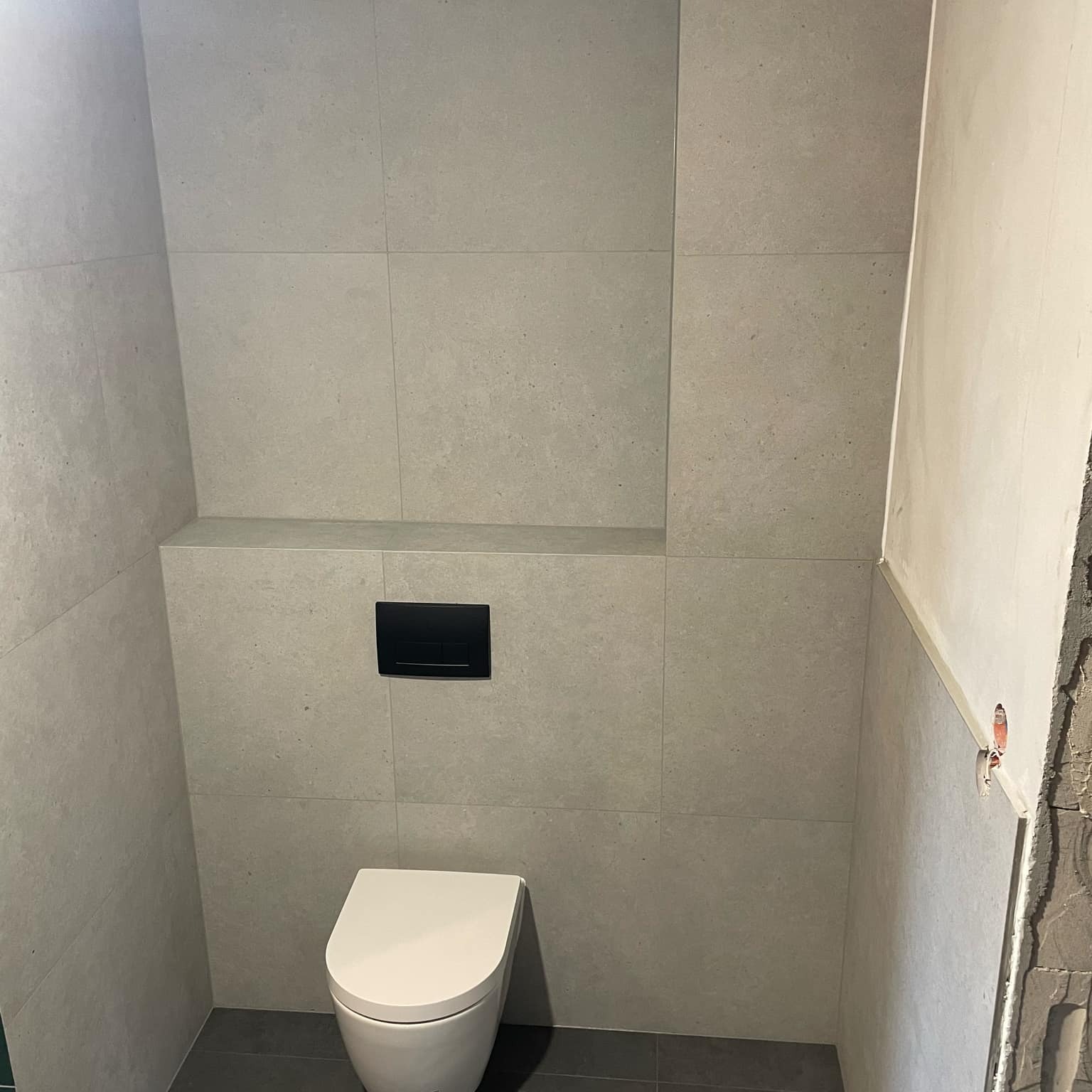 Świeżo wykafelkowana ściana łazienki z zamontowaną podtynkową spłuczką i wiszącą toaletą, widoczne niedokończone wykończenie ściany po prawej stronie.