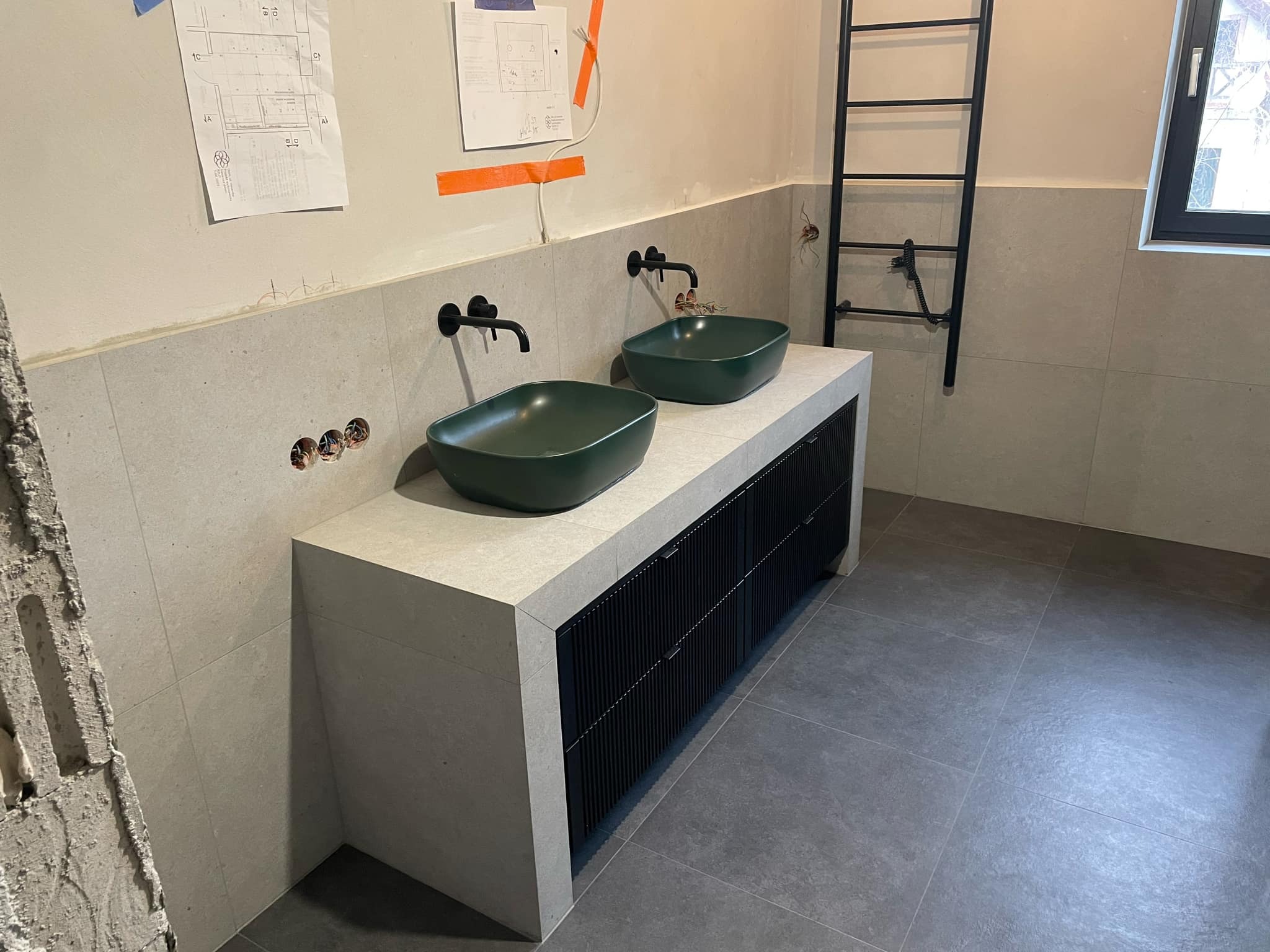 Łazienka w trakcie remontu: Dwie zielone, kwadratowe umywalki nablatowe z czarną armaturą na blacie z szarej płytki, czarne szafki pod umywalkami, szare płytki na ścianach i podłodze, widoczne...