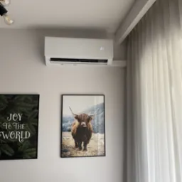 Biała klimatyzacja ścienna w nowoczesnym wnętrzu, obok wiszą grafiki, w tym obraz z krową szkocką. Zasłony w oknie, minimalistyczny design.