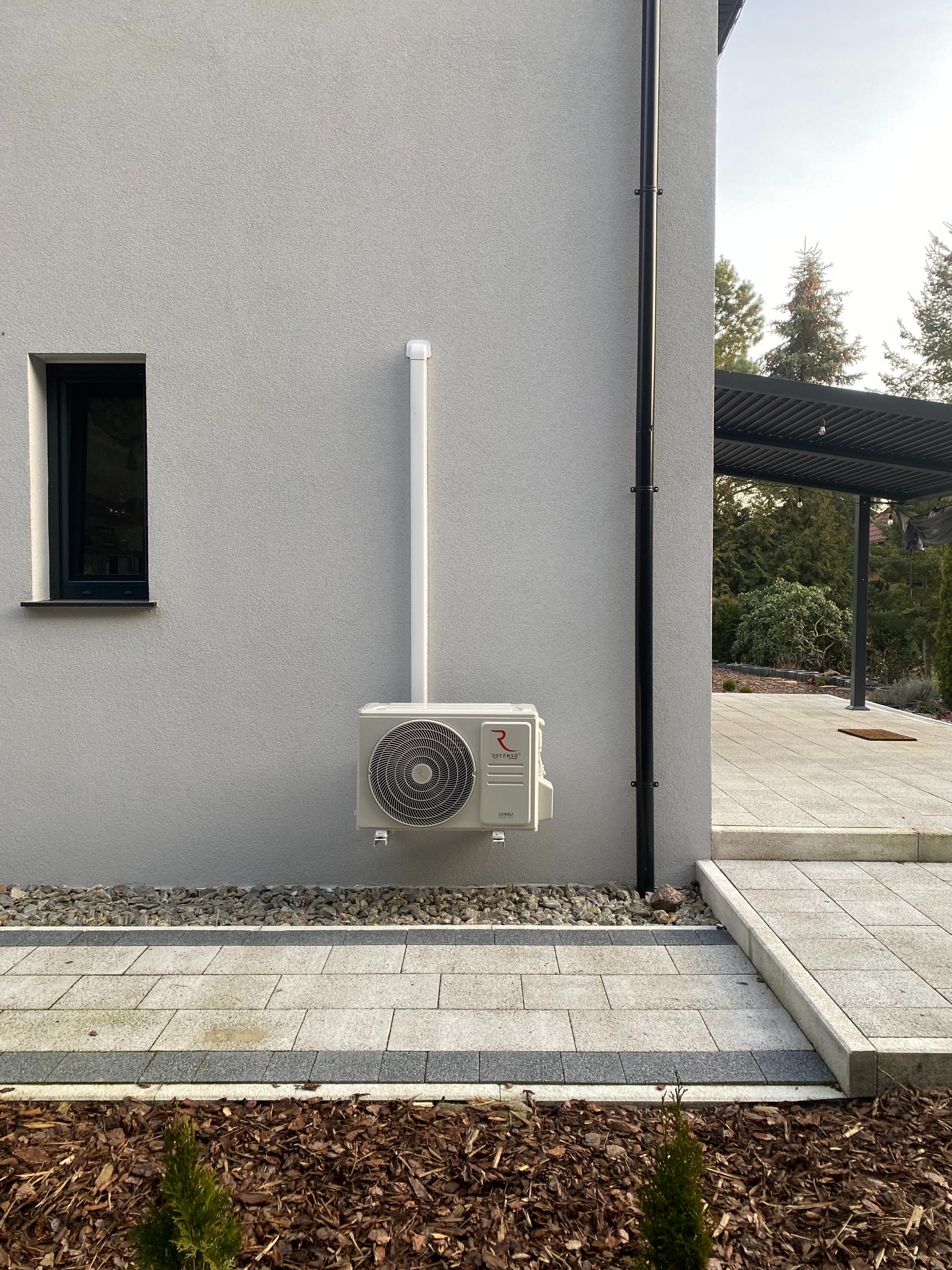 Biały klimatyzator Rotenso zamontowany na elewacji domu, z widoczną instalacją odprowadzającą. Obok okno i rynna, w tle zadaszenie tarasu i drzewa.