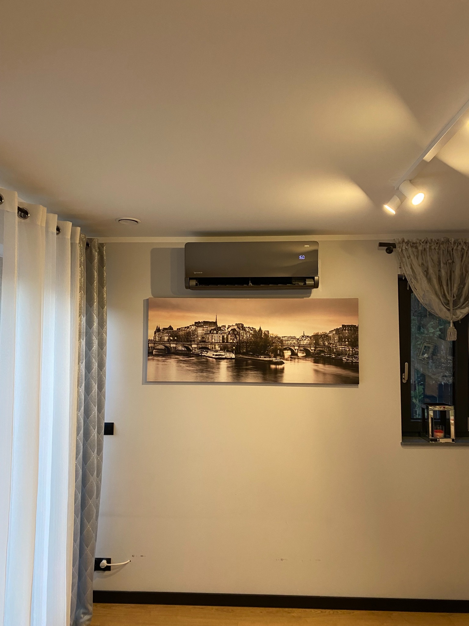 Szara klimatyzacja Rotenso zamontowana nad obrazem z motywem miejskim w salonie. Widoczne zasłony i oświetlenie punktowe.