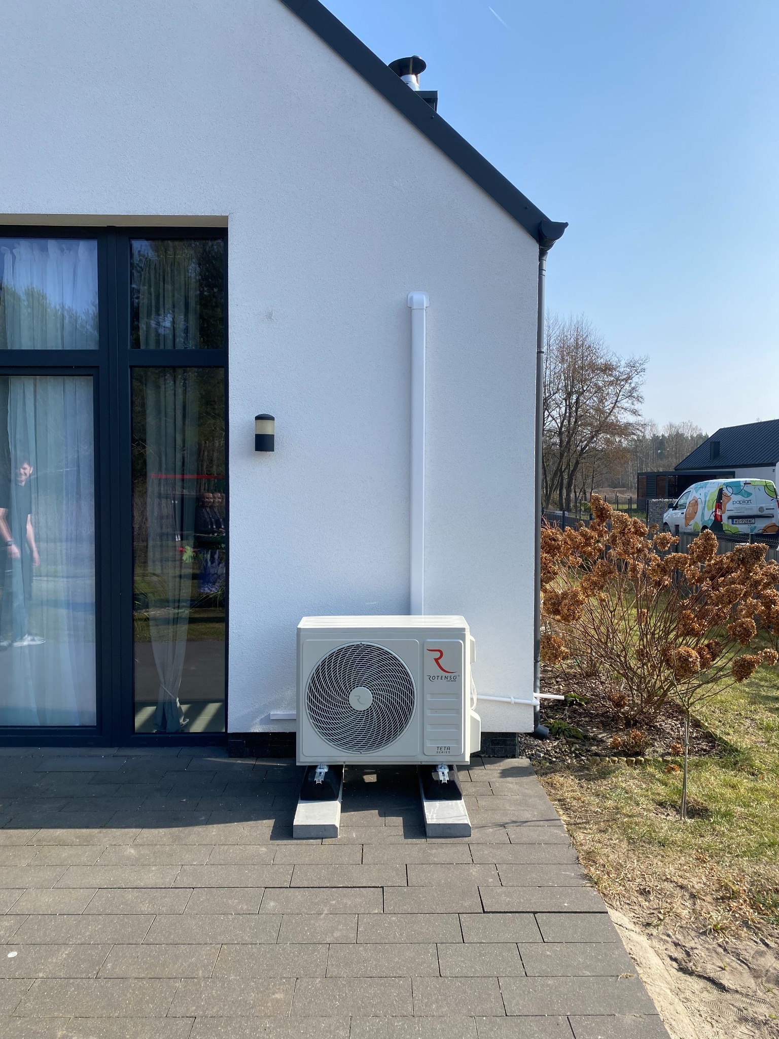 Biała jednostka zewnętrzna klimatyzacji Rotenso Teta zamontowana na szarym podłożu przy ścianie domu z czarnymi oknami i rurą odprowadzającą.