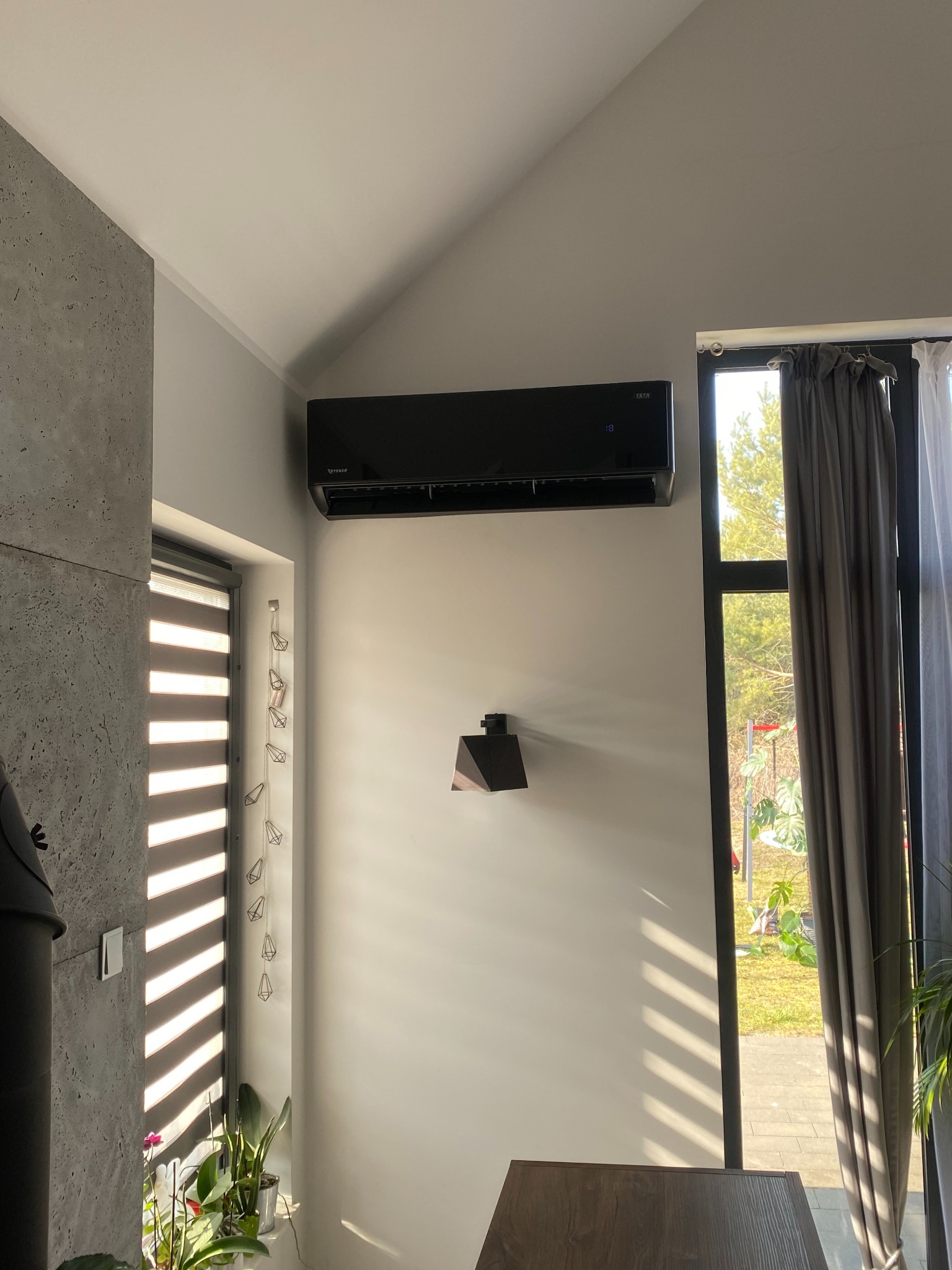 Czarny klimatyzator marki Rotenso zamontowany na białej ścianie w nowoczesnym salonie z oknem i zasłonami, oświetlonym promieniami słońca.
