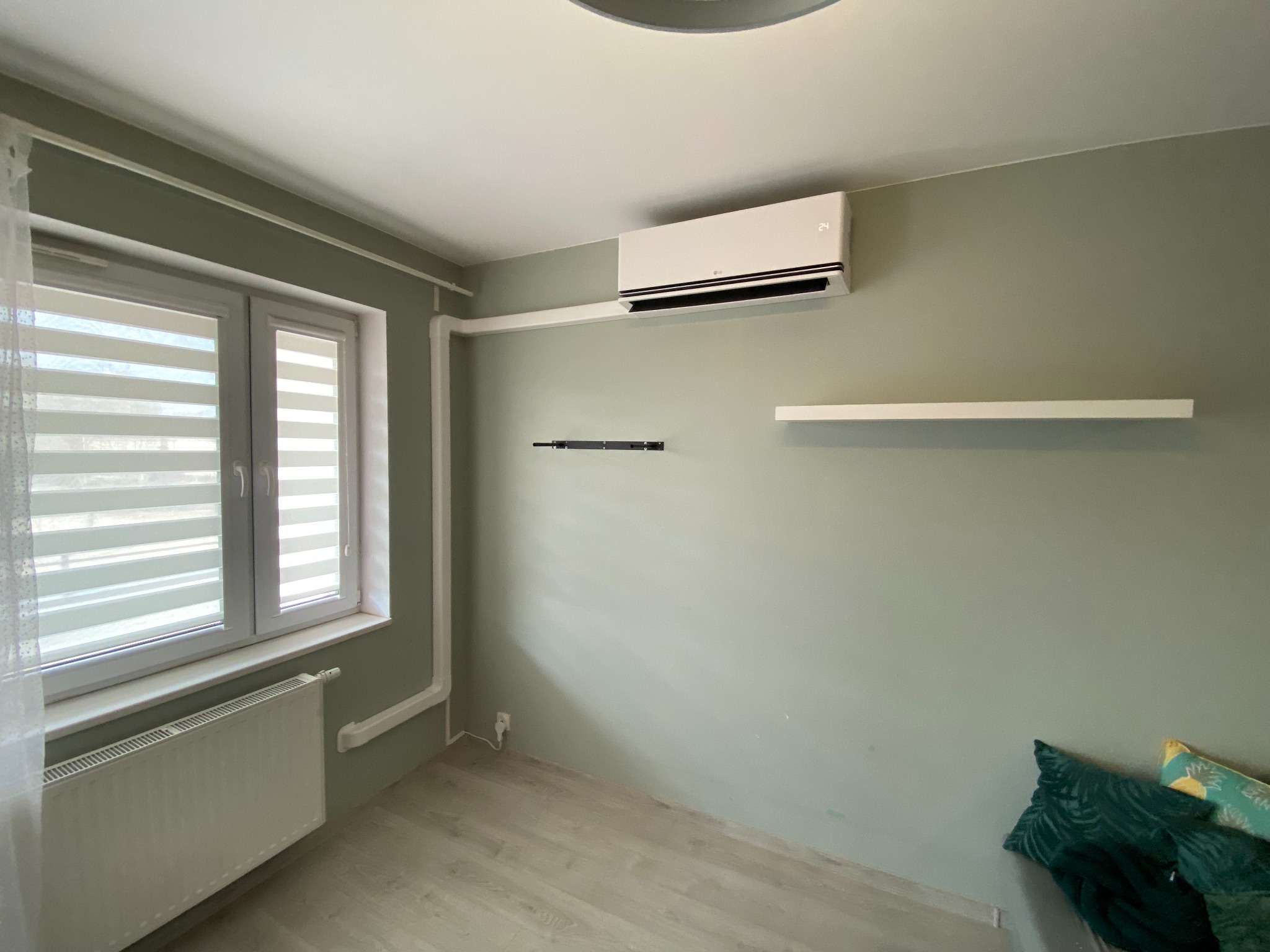 Klimatyzator LG na ścianie w pokoju o zielonych ścianach, z grzejnikiem pod oknem i drewnianą podłogą. Widoczna instalacja rur i półka ścienna.