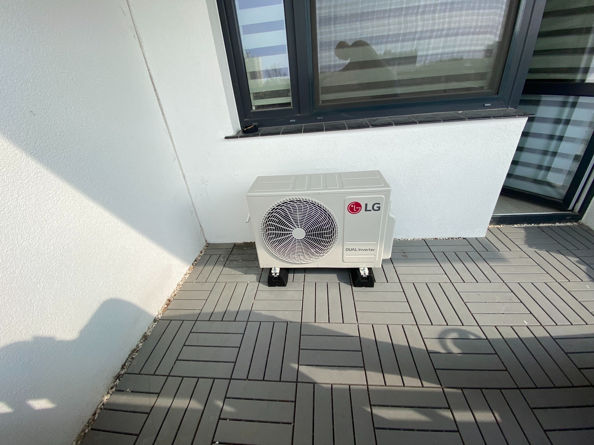 Zewnętrzna jednostka klimatyzacji LG Dual Inverter zamontowana na balkonie z szarymi płytkami podłogowymi, widoczna pod oknem z żaluzjami.