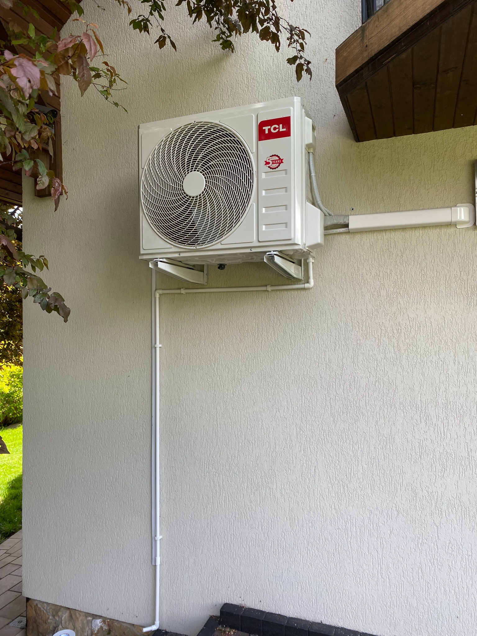 Biała jednostka zewnętrzna klimatyzacji TCL zamontowana na elewacji budynku z widocznymi rurami odprowadzającymi. Widok z dołu, fragment roślinności w tle.