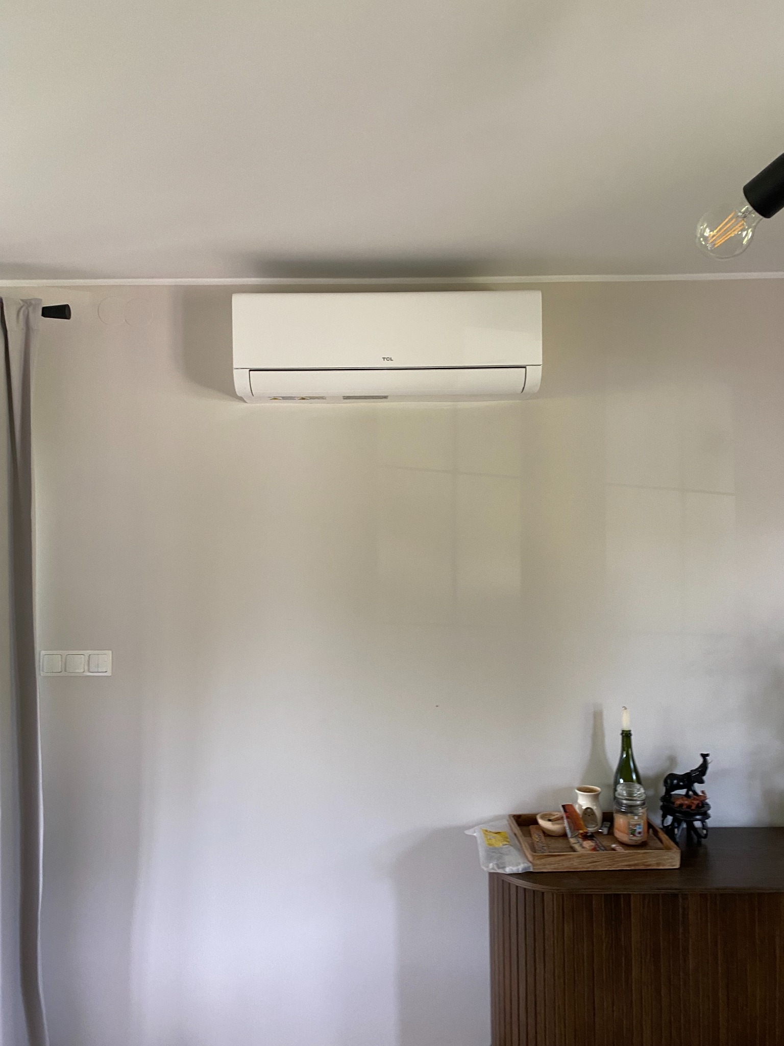 Biały klimatyzator TCL zamontowany na ścianie w pokoju z drewnianą komodą, na której stoją dekoracje. Widoczne oświetlenie sufitowe i zasłona okienna.