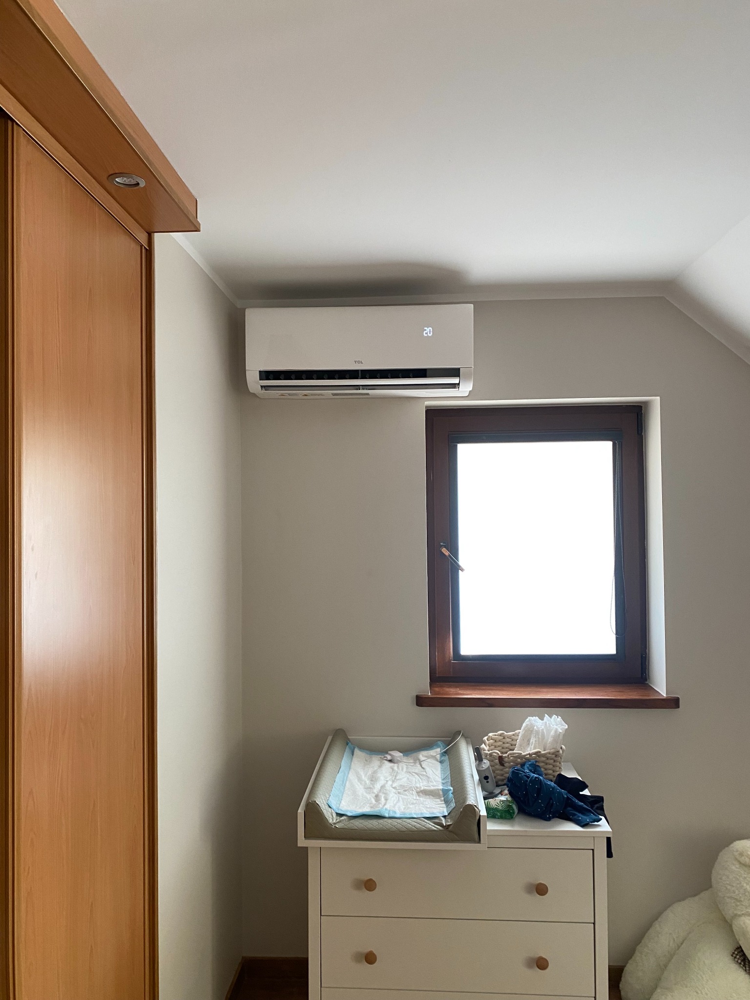 Biała klimatyzacja TCL zamontowana nad oknem w pokoju dziecięcym z komodą i przewijakiem. Widoczna szafa z jasnego drewna.