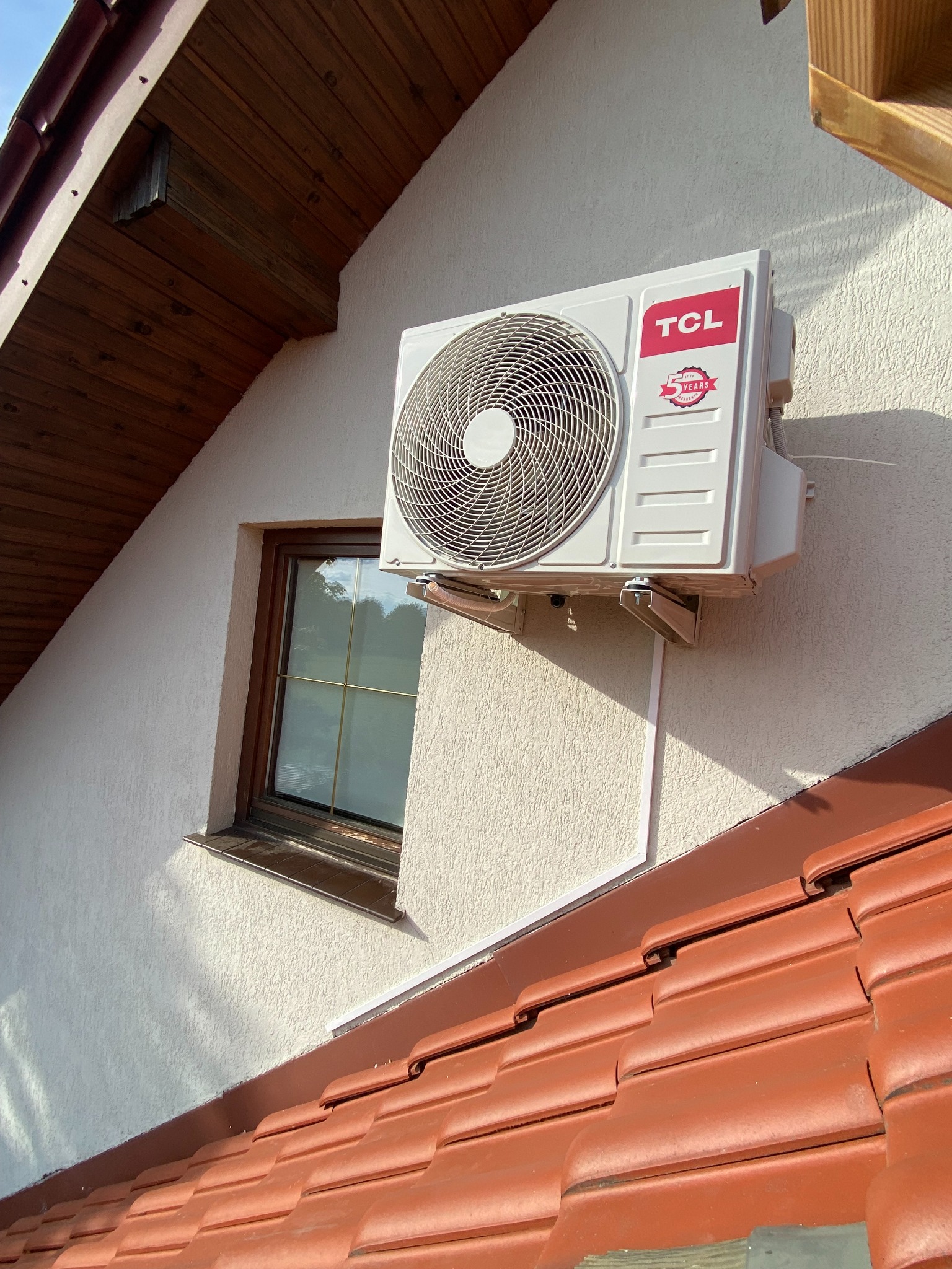 Biała klimatyzacja TCL zamontowana na elewacji domu obok okna, widoczna z perspektywy dachu z czerwoną dachówką. Oświetlenie słoneczne.