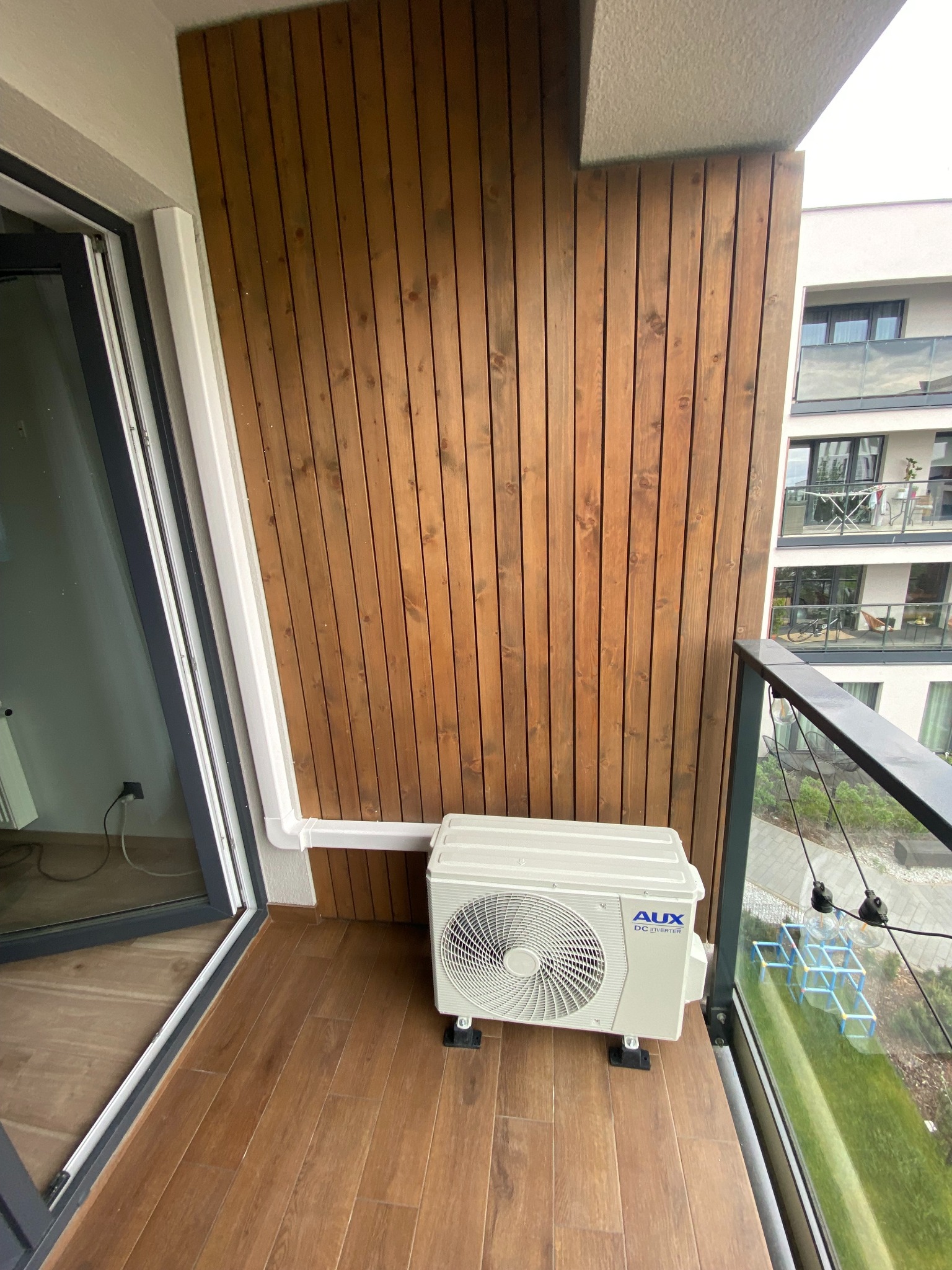Biała jednostka zewnętrzna klimatyzacji AUX zamontowana na balkonie z drewnianą podłogą i ścianką. Widok na sąsiedni budynek przez szklaną balustradę.