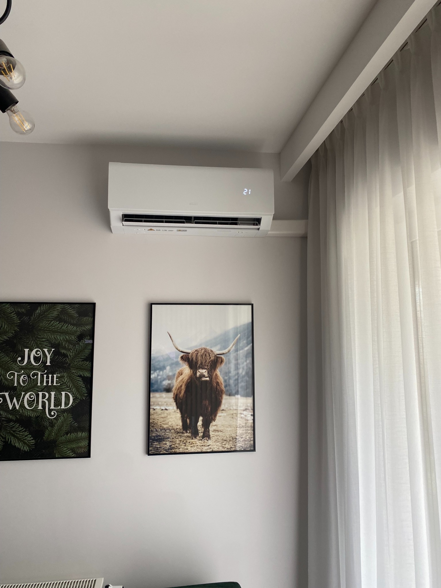 Biała klimatyzacja ścienna w nowoczesnym wnętrzu, obok wiszą grafiki, w tym obraz z krową szkocką. Zasłony w oknie, minimalistyczny design.