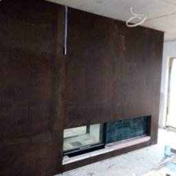 Flismarbud - Montaż ściany z płyty karton gips z wnęką na kominek z wykończeniem z płytek ceramicznych, widoczne kable pod sufitem i surowe wykończenie pomieszczenia.